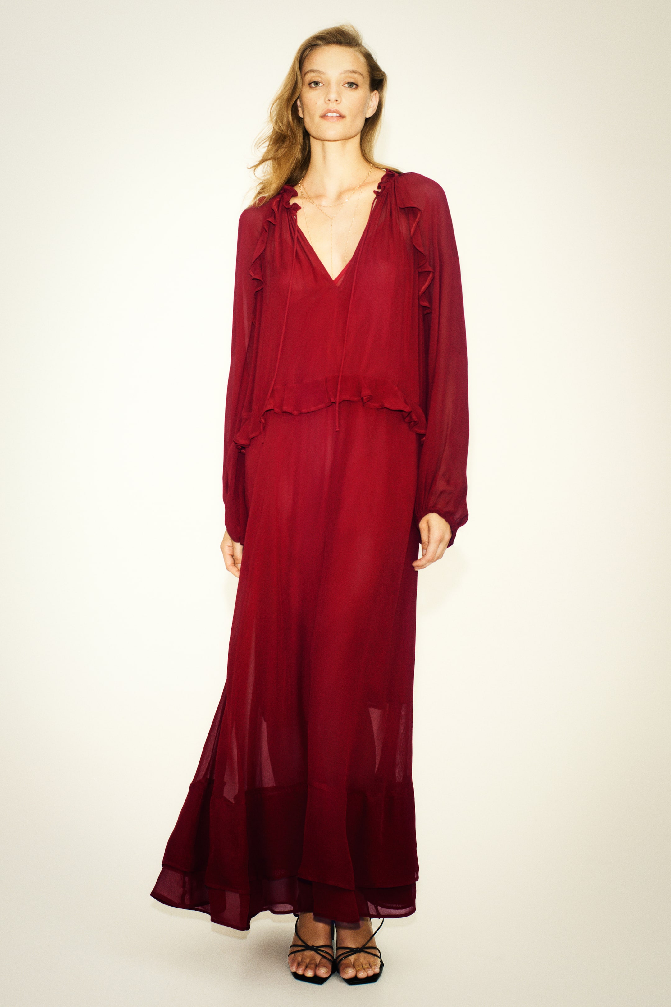 View larger image: A front-facing view shows a dark red maxi dress crafted from semi-sheer fabric, featuring a deep V-neckline with thin ties and ruffled flounce details along the shoulders and waist. The dress is styled with long balloon sleeves and a gently flared skirt that showcases tiered ruffles at the hem.