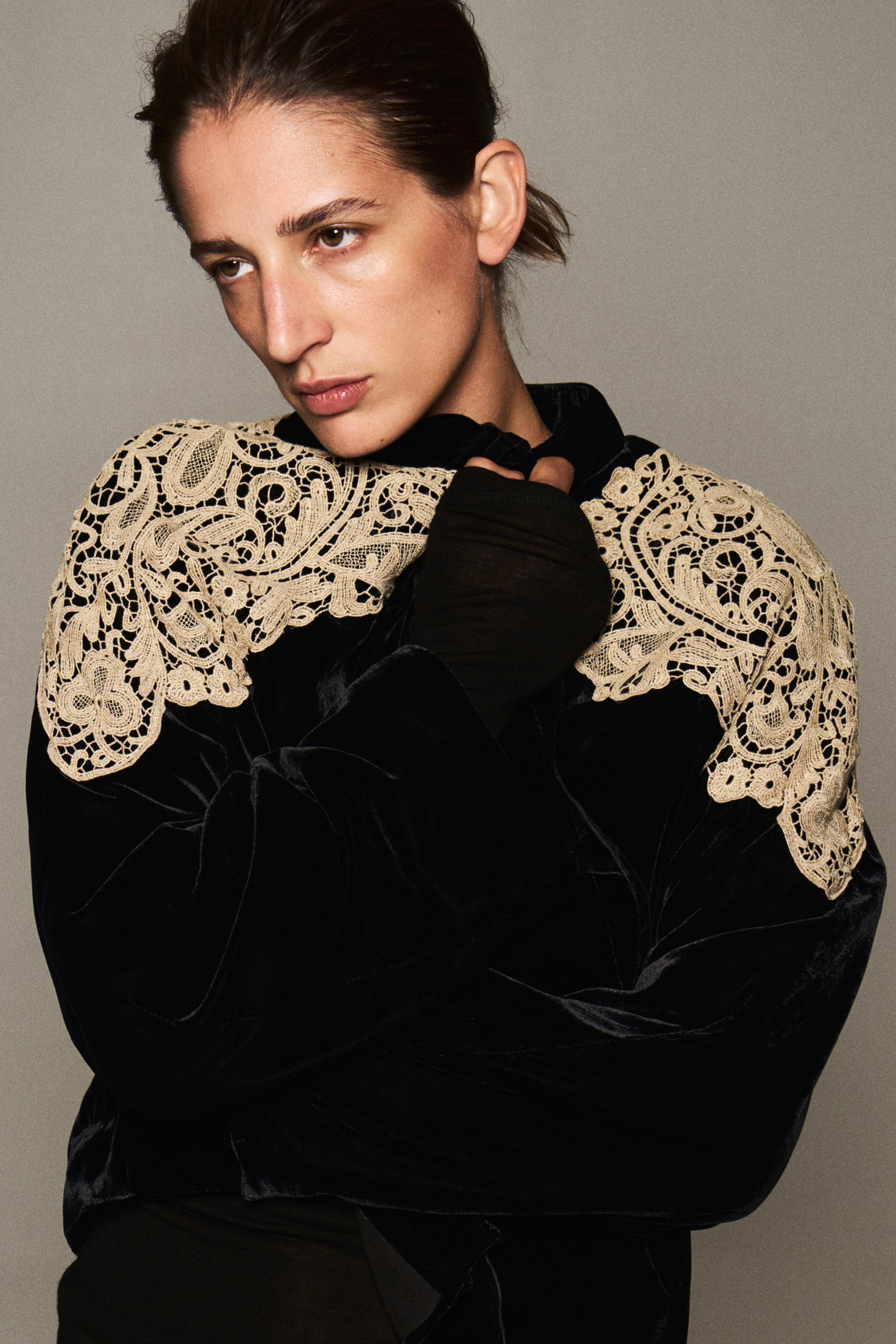 View larger image: A black velvet jacket featuring intricate cream-coloured lace overlays on the shoulders and a high band collar is worn by the model. The model holds a gloved hand near the collar, highlighting the contrast between the dark velvet fabric and the light lace detail.