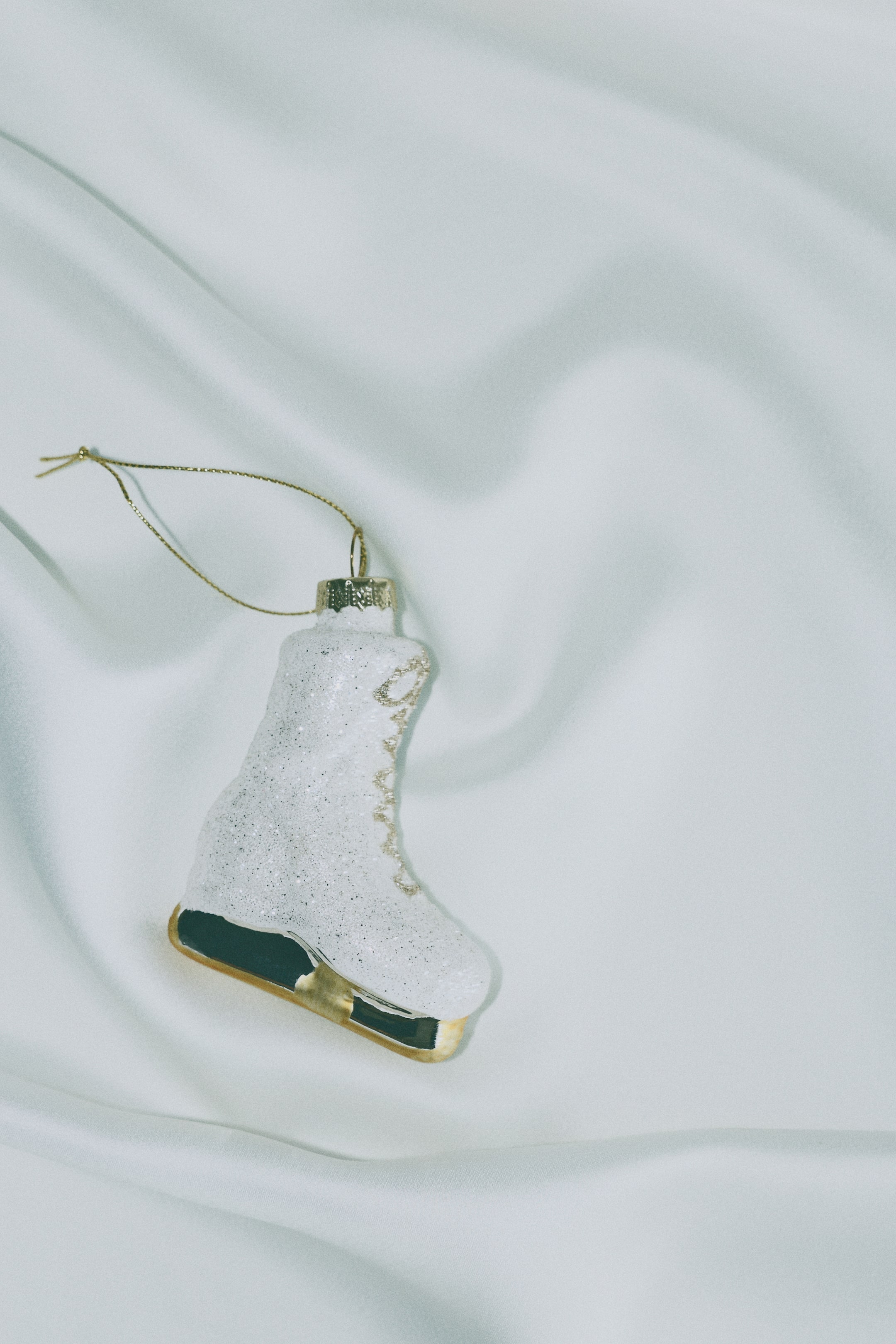 Visualizza immagine più grande: Una decorazione natalizia in vetro a forma di pattino da ghiaccio è mostrata di profilo, con uno stivale bianco glitterato, lacci dipinti in oro e una lama color oro. Un cappuccio in metallo dorato è fissato sulla parte superiore dello stivale e sostiene un sottile anello di cordino dorato per appenderla.