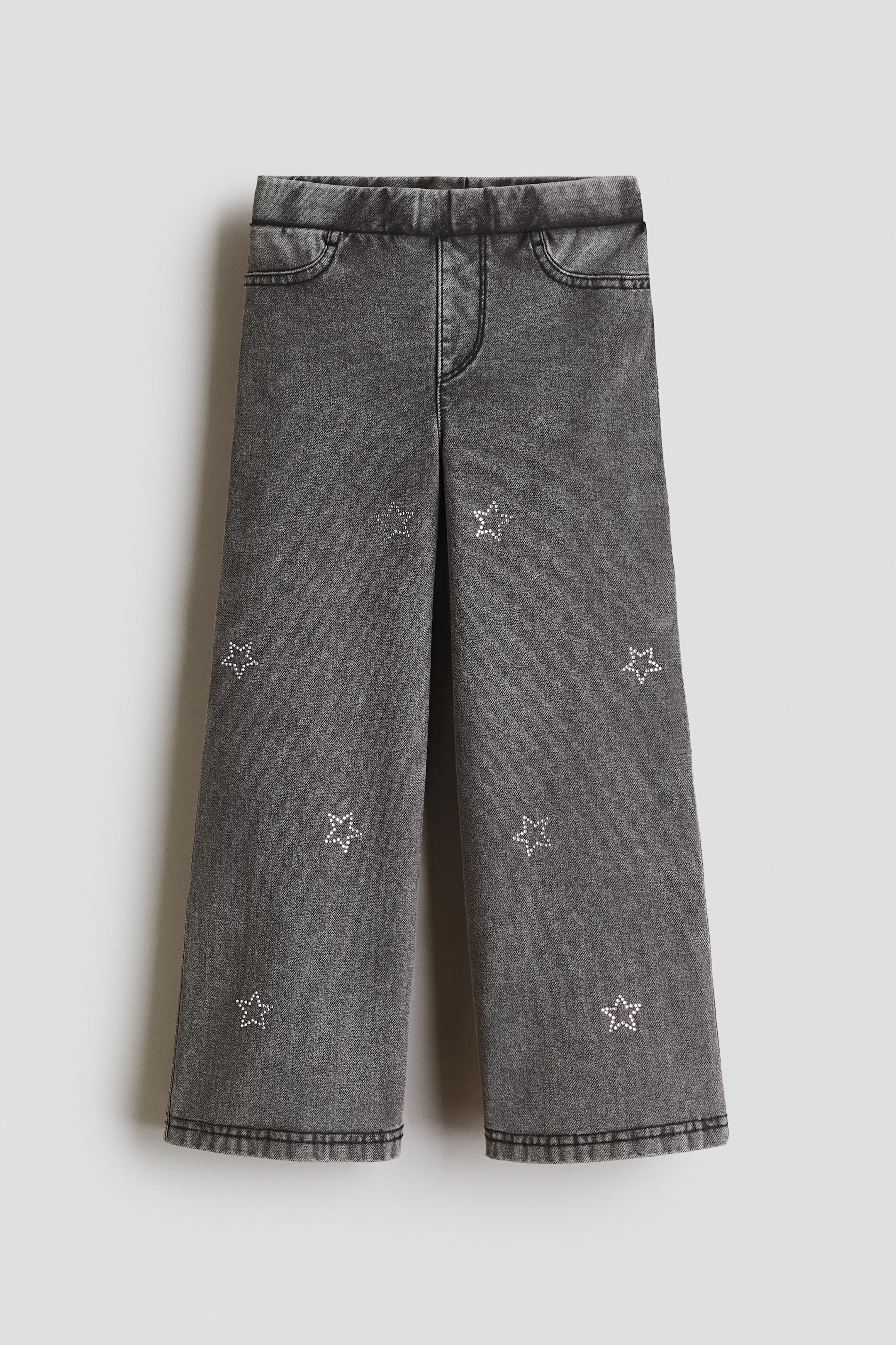 Grotere afbeelding bekijken: Van voren gezien zijn deze jeggings uitgevoerd in een zware grijze jersey met denimlook, met een verstelbare elastische tailleband, een nepgulp en decoratieve zakken aan de voorkant, terwijl sprankelende stermotieven de pijpen sieren.
