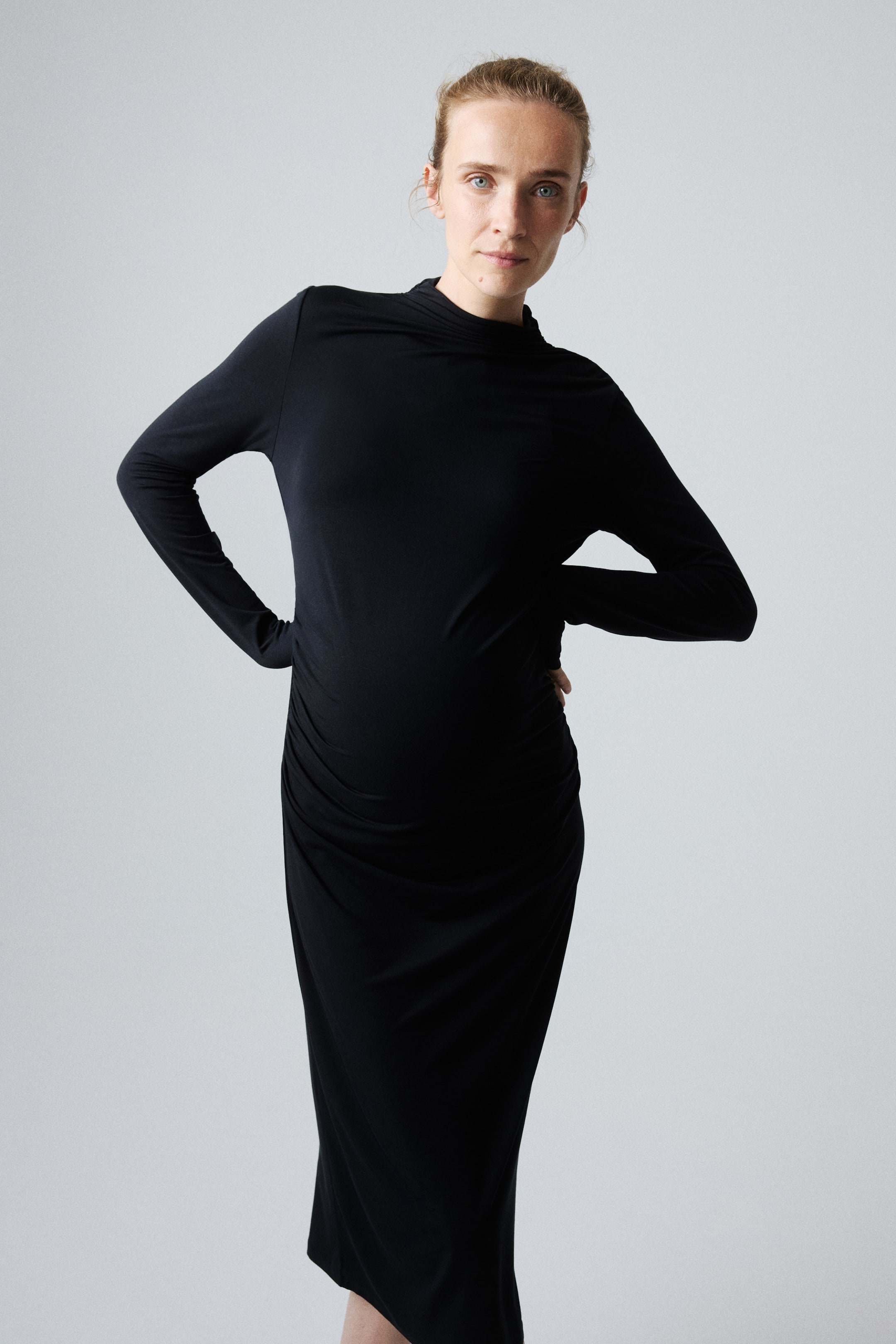 Grösseres Bild ansehen: Ein schwarzes Midi-Kleid mit hohem Kragen, langen Ärmeln und Raffung an einer Seite des Rocks wird von dem Model getragen. Das Model hat die Hände in die Hüften gestützt.