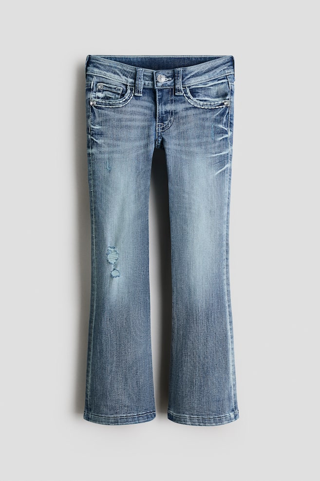 Jeans in gewassen denim tonen een vooraanzicht, met een lage taille en een ritssluiting met knoopsluiting, samen met voorzakken en een muntzakje. Ze vertonen versleten details, waaronder lichte plooien op de dijen en een versleten plek op het linkerbeen, en zijn ontworpen met een skinny fit van de taille tot de dij, uitlopend in een bootcut pijp van de knie tot de zoom. - 1