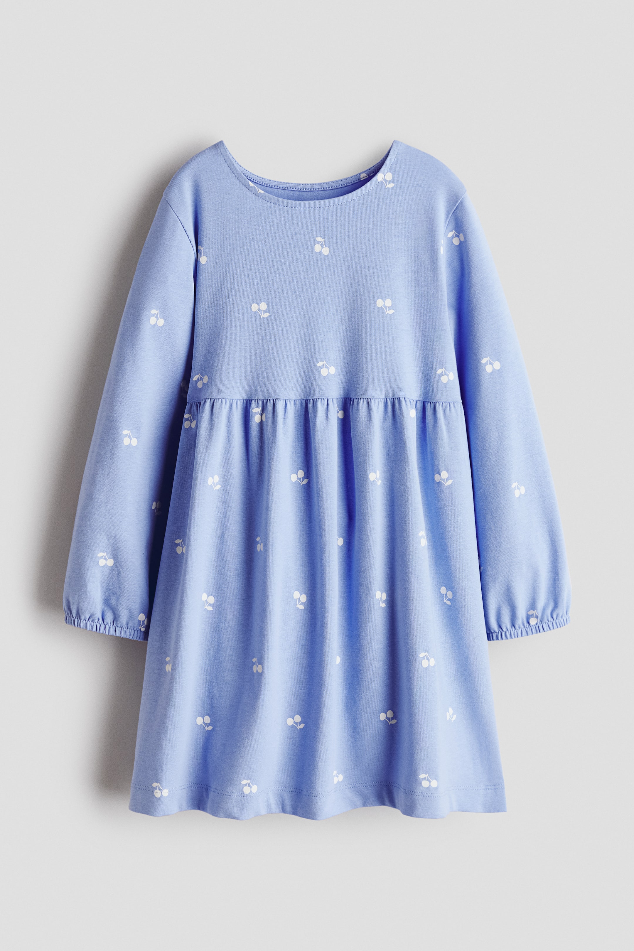 View larger image: A light blue jersey dress features an all-over white cherry print and is shown from the front. It has a round neckline, long sleeves with elastic cuffs, and a gathered seam at the waist.