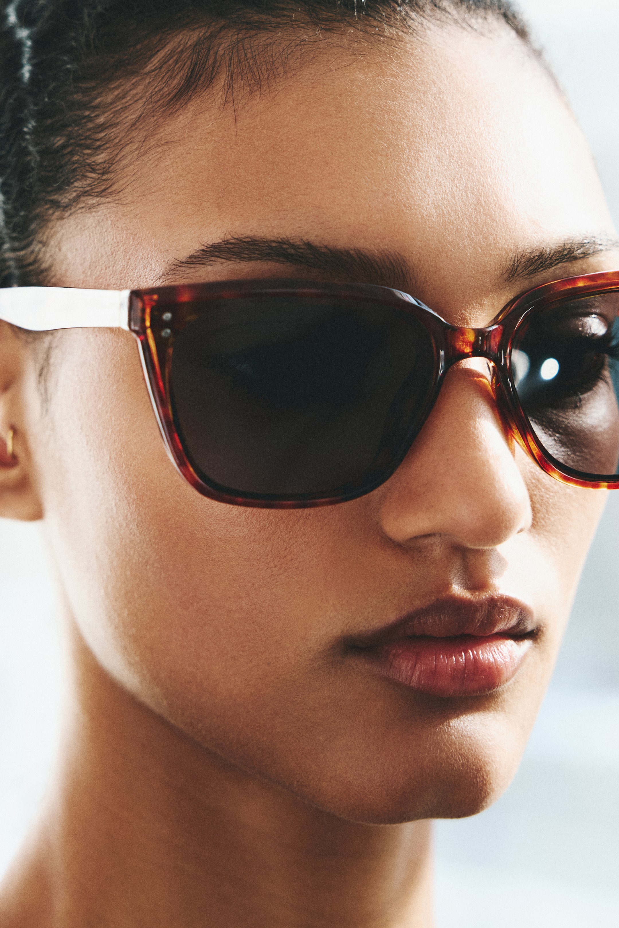 Größeres Bild ansehen: Tortoise-Sonnenbrillen mit dunklen getönten Gläsern in rechteckigen Fassungen, die kleine Metallakzente an den äußeren Ecken aufweisen. Die Fassungen kontrastieren mit weißen Bügeln, und die Sonnenbrillen werden leicht nach rechts geneigt getragen.