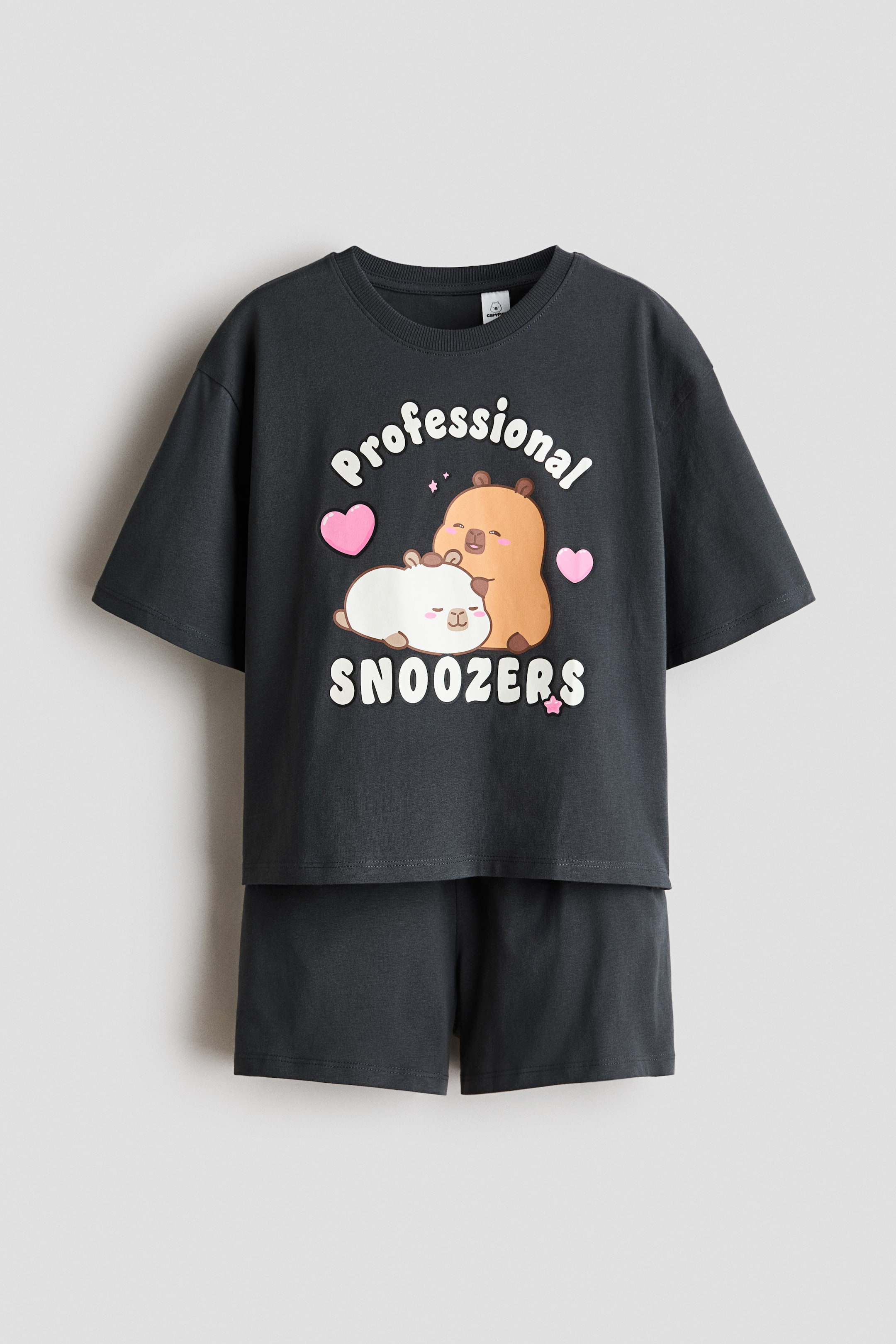 Visualizza immagine più grande: Un set da pigiama composto da una maglietta e pantaloncini di colore grigio scuro è esposto frontalmente. La maglietta presenta un girocollo e maniche corte, con una stampa grafica bianca che riporta "Professional SNOOZERS" accanto a due animali dei cartoni animati addormentati, cuori rosa e stelle. I pantaloncini coordinati grigio scuro includono una vita elasticizzata.