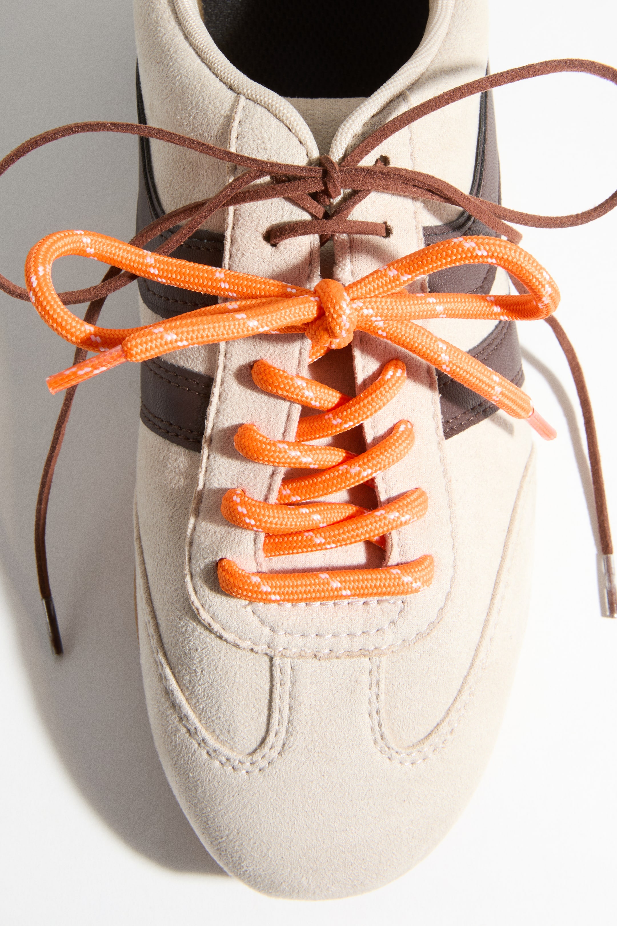 Voir une image plus grande: Une chaussure de sport en daim beige clair est présentée vue de dessus, avec des bandes latérales marron foncé et deux ensembles de lacets. Les œillets inférieurs sont enfilés avec des lacets orange vif, mouchetés, noués en un nœud proéminent, tandis que des lacets plus fins, marron foncé, sont noués au-dessus.