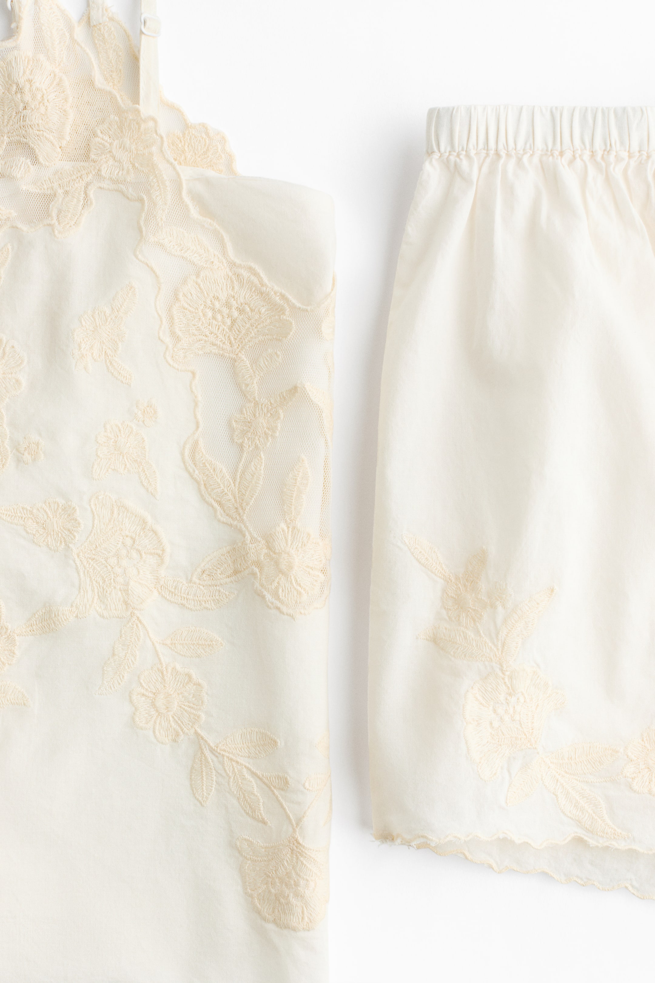 Ver imagen más grande: Un conjunto de ropa de dos piezas en color marfil claro que incluye un top de camisola y pantalones cortos a juego. La camisola presenta un delicado bordado floral sobre malla transparente con un escote festoneado y tirantes finos, mientras que los pantalones cortos tienen una cintura elástica con bordado floral coordinado cerca del dobladillo festoneado.