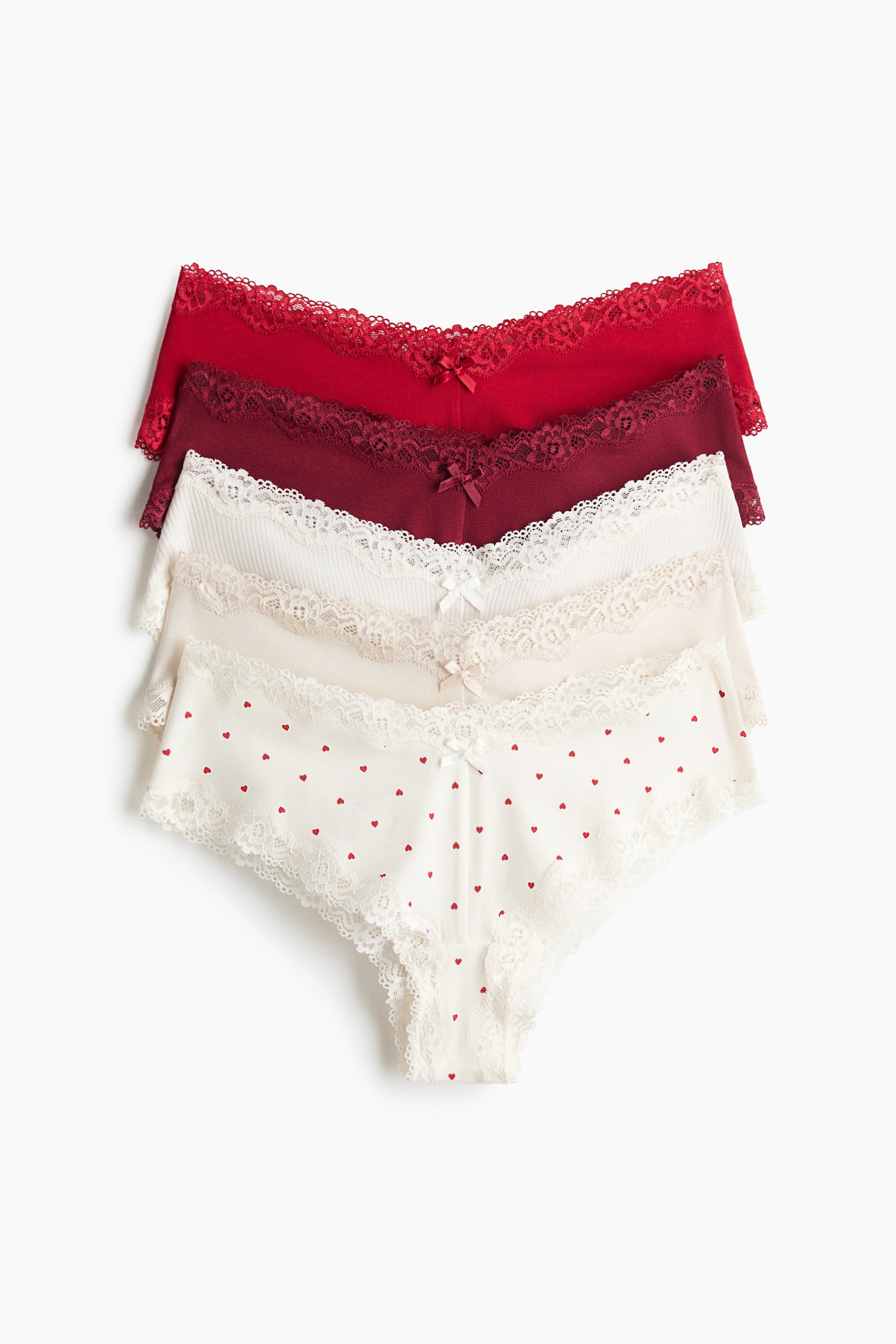 View larger image: Five pairs of low-rise briefs are arranged in a stacked, front-facing display. The assortment features solid red, burgundy, ribbed off-white, beige, and heart-patterned styles, with each pair showcasing delicate lace trims and a small bow at the centre of the V-shaped waistband.