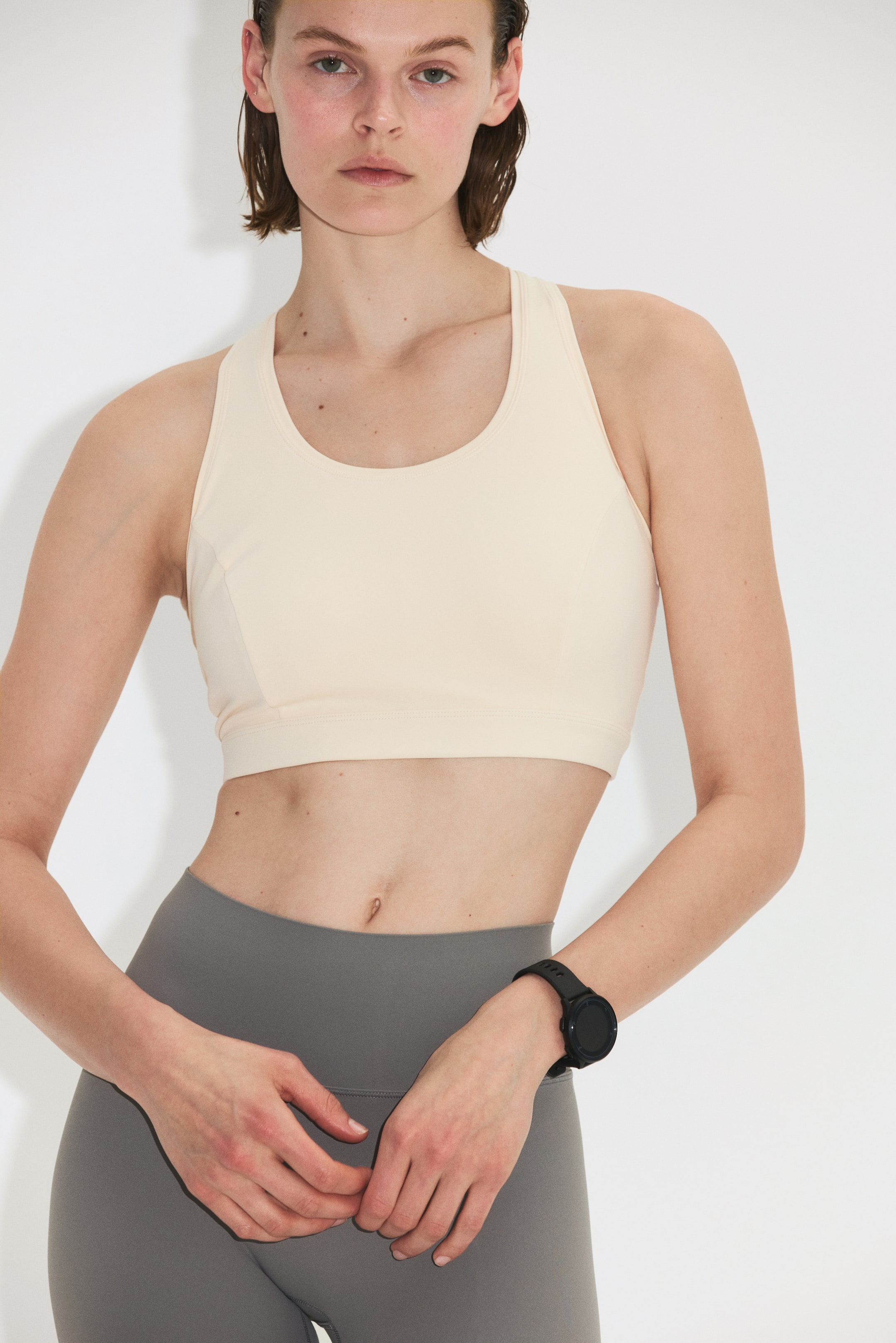 Ver imagem maior: Um sutiã desportivo bege claro, com decote redondo, alças largas, painéis laterais subtis e uma faixa elástica larga na bainha, está a ser usado pela modelo. A modelo também está a usar leggings cinzentas.