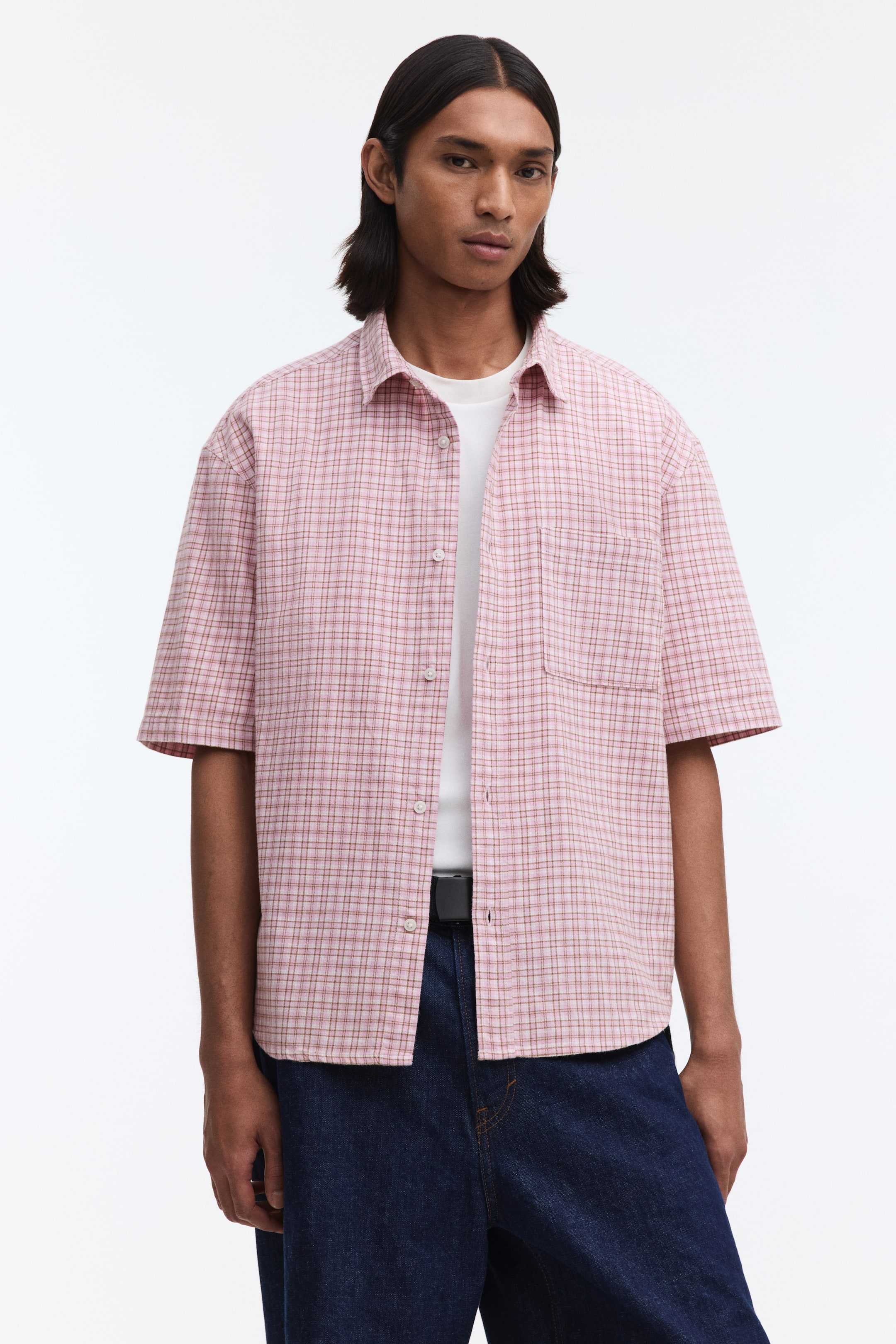 View larger image: A pink plaid short-sleeved shirt menampilkan kolar leper dan butang putih di bahagian hadapan. Corak plaid ini memaparkan garis-garis merah jambu muda, merah jambu gelap, dan coklat nipis, dengan satu poket tampal kelihatan di dada kiri. Baju ini dipakai terbuka di atas t-shirt putih dan sebahagiannya diselitkan ke dalam jeans biru gelap, memberikan kesan potongan santai.