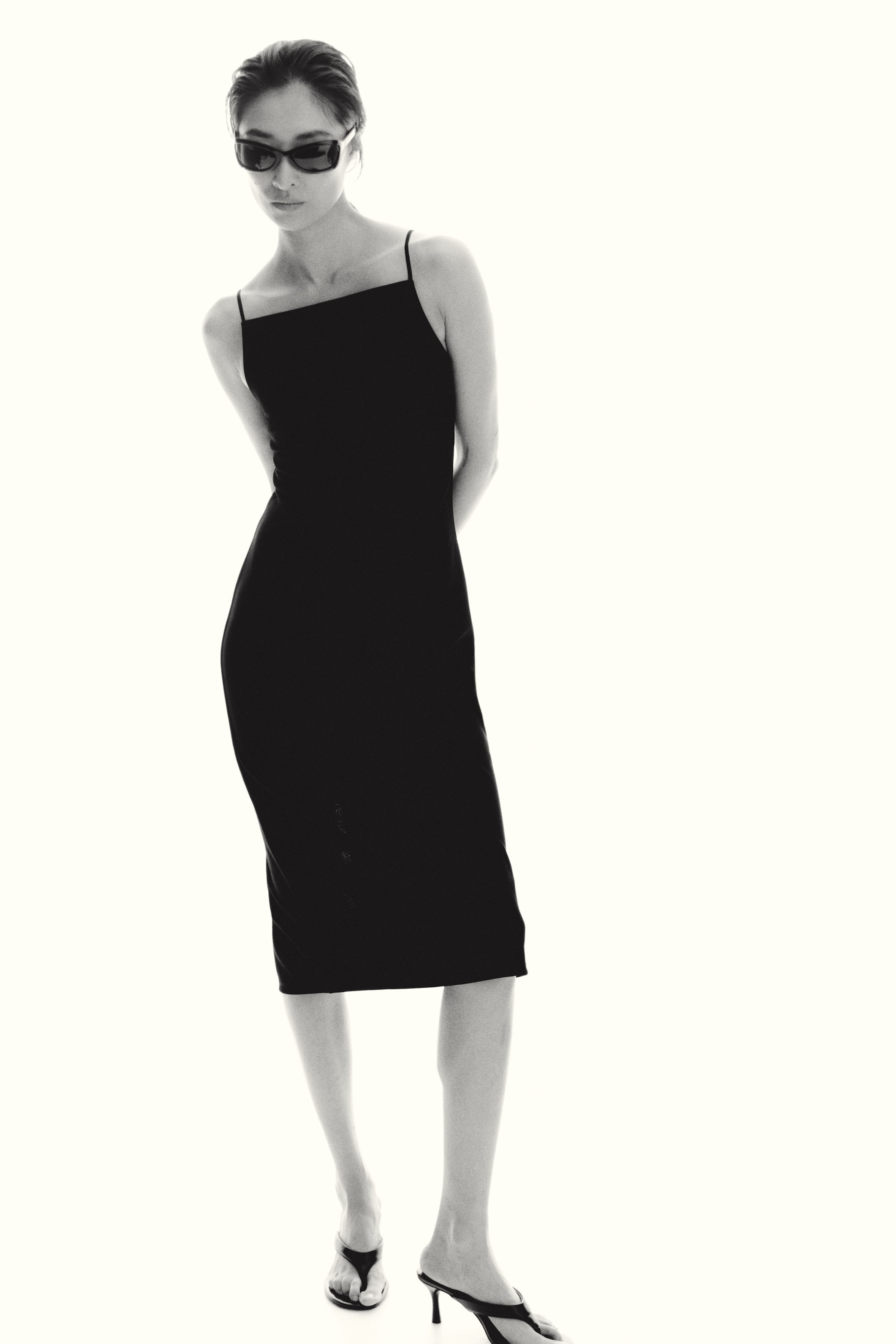 View larger image: The model is wearing a black, calf-length fitted dress featuring a straight neckline and narrow shoulder straps. She stands poised, elegantly highlighting the dress.