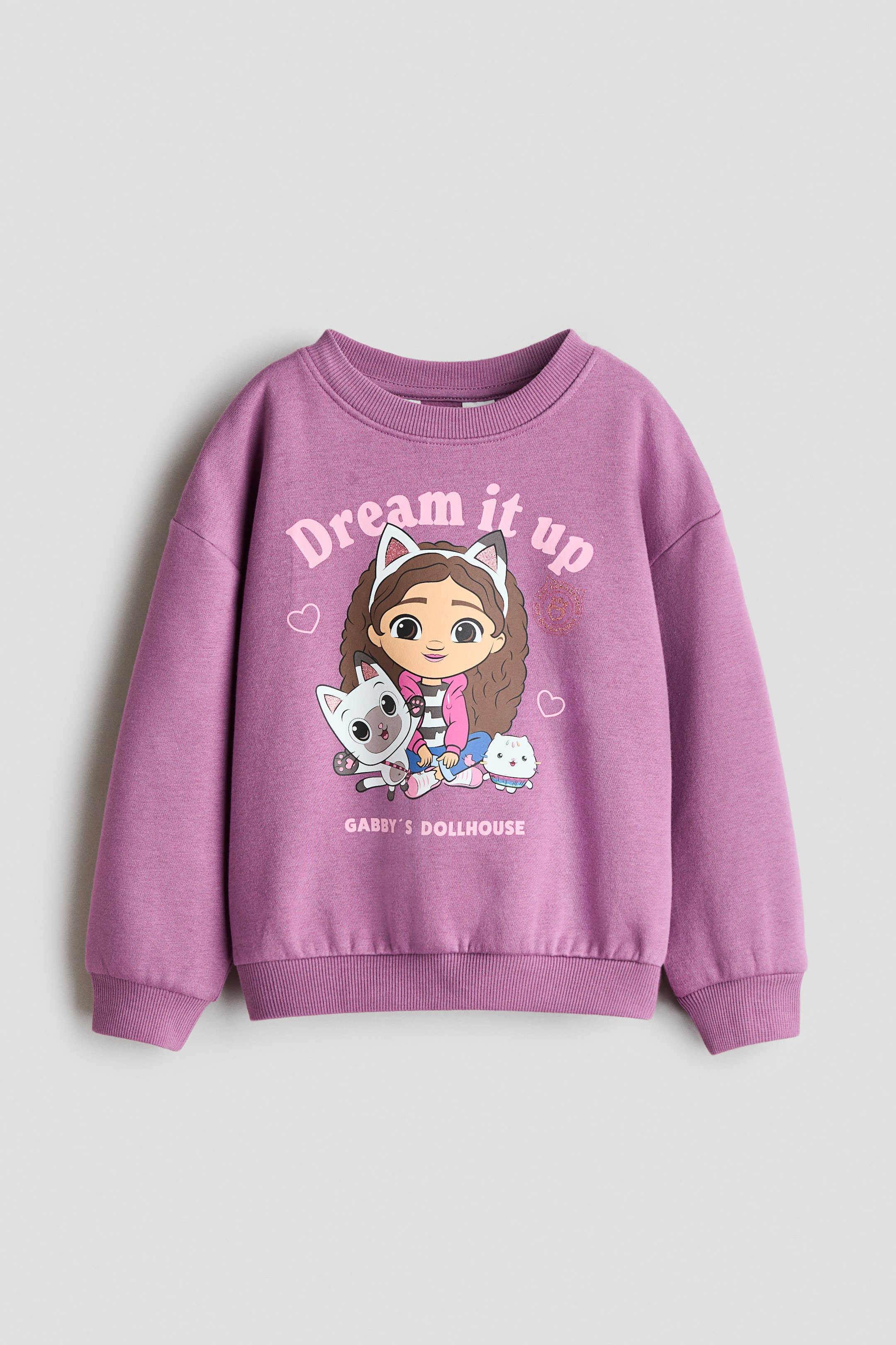 View larger image: A purple sweatshirt is shown front-facing, featuring a round, rib-trimmed neckline, dropped shoulders, and long sleeves with ribbed cuffs. The hem is also ribbed, and the front features a large graphic print of a cartoon girl with cat ears and two cats, along with the text "Dream it up" and "Gabby's Dollhouse."