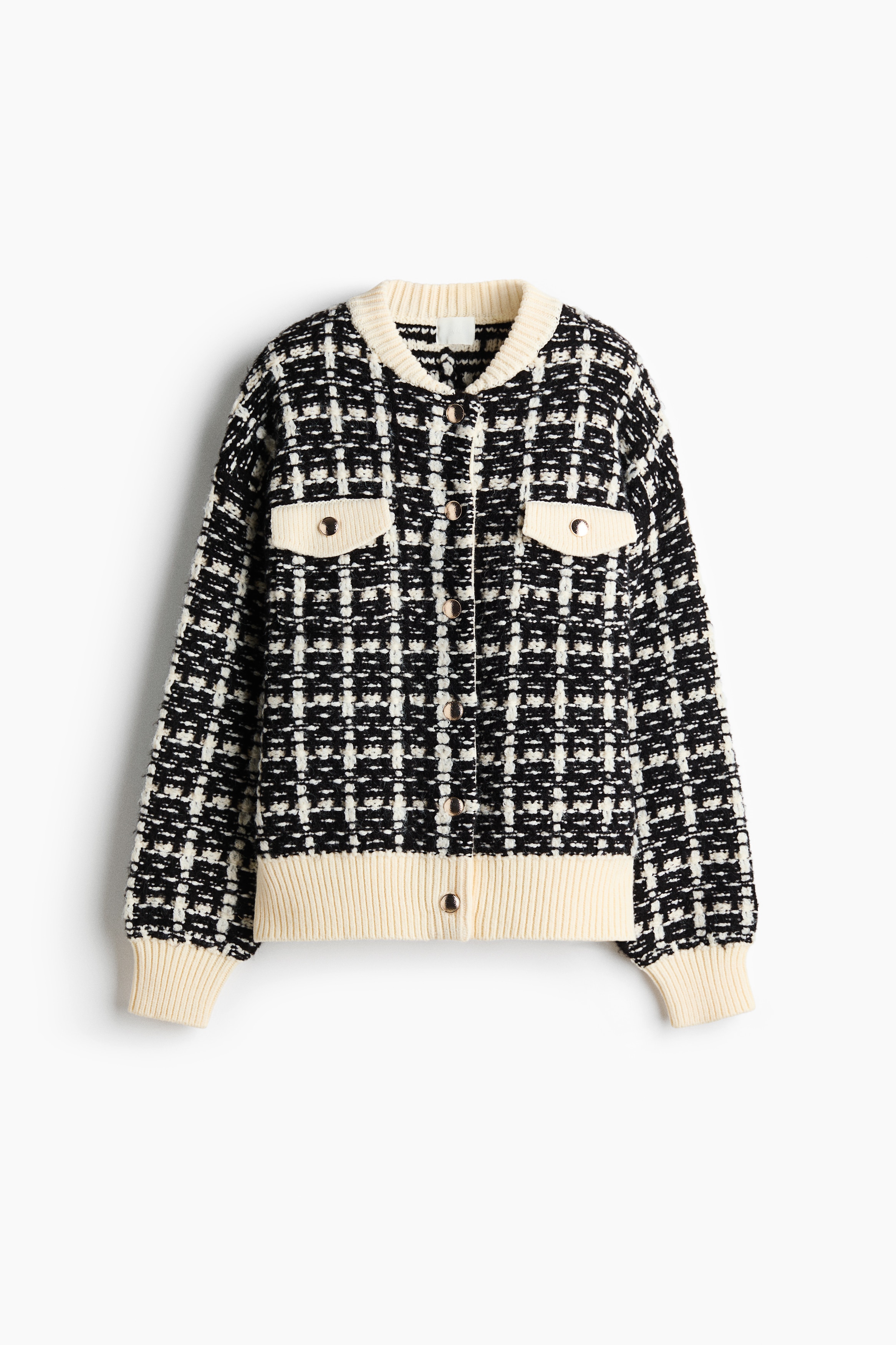La modelo lleva una chaqueta de tejido texturizado en blanco y negro sobre un top blanco y jeans negros. La chaqueta presenta un patrÃ³n de cuadros, ribete acanalado blanco en el cuello, los puÃ±os y el dobladillo, y bolsillos con tapa abotonada en el pecho.