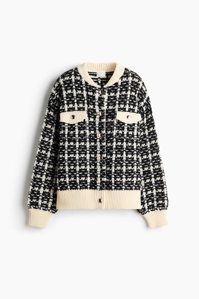 A black and white tweed-patterned knit cardigan is facing front, displaying a button-up front closure with gold-toned buttons. It is featuring a contrasting cream-colored ribbed crew neck, hem, and cuffs, along with two chest flap pockets adorned with matching gold buttons. - 1