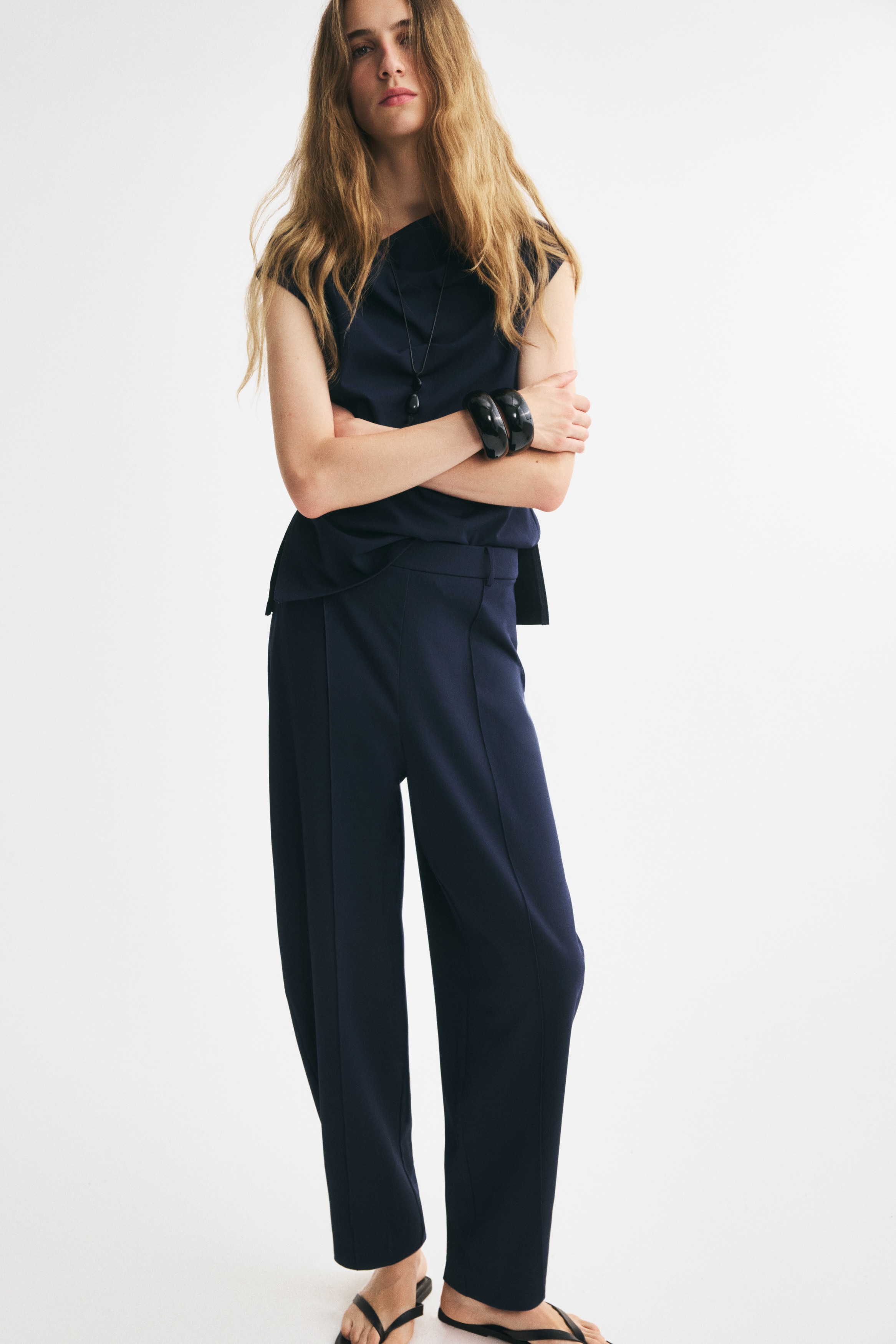 Donkerblauwe broek toont een vooraanzicht, met een platte tailleband met riemlussen en gestikte voorkreukels. De broek heeft een barrel-leg pasvorm, die taps toeloopt naar de enkels met een nette zoom.