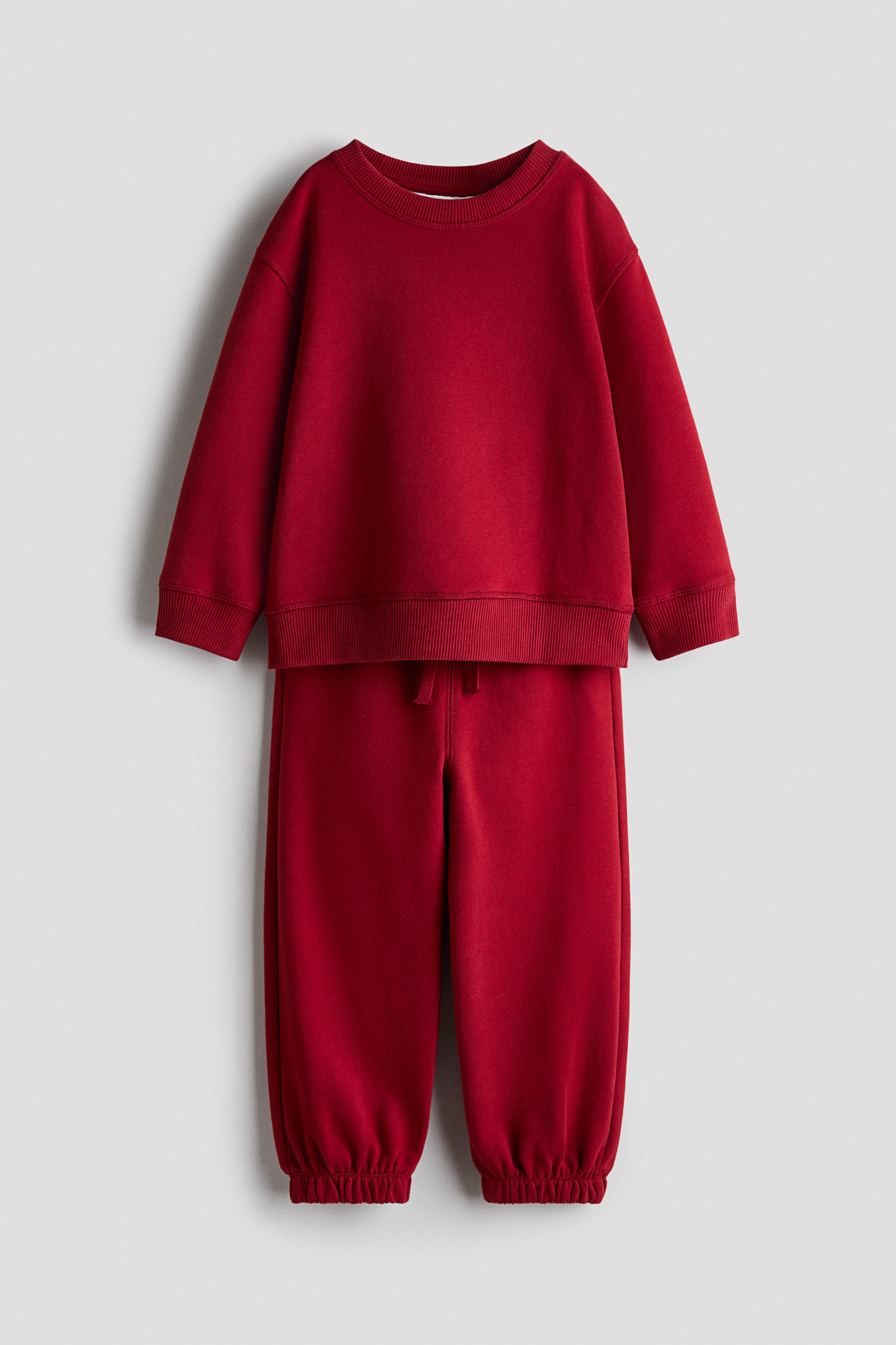 A child sitting cross-legged and facing forward is wearing a red cotton sweat set featuring a sweater with a round ribbed neckline and wide ribbed cuffs. The sweater is layered over a white collared shirt, with the collar and cuffs visible, paired with matching red track pants.