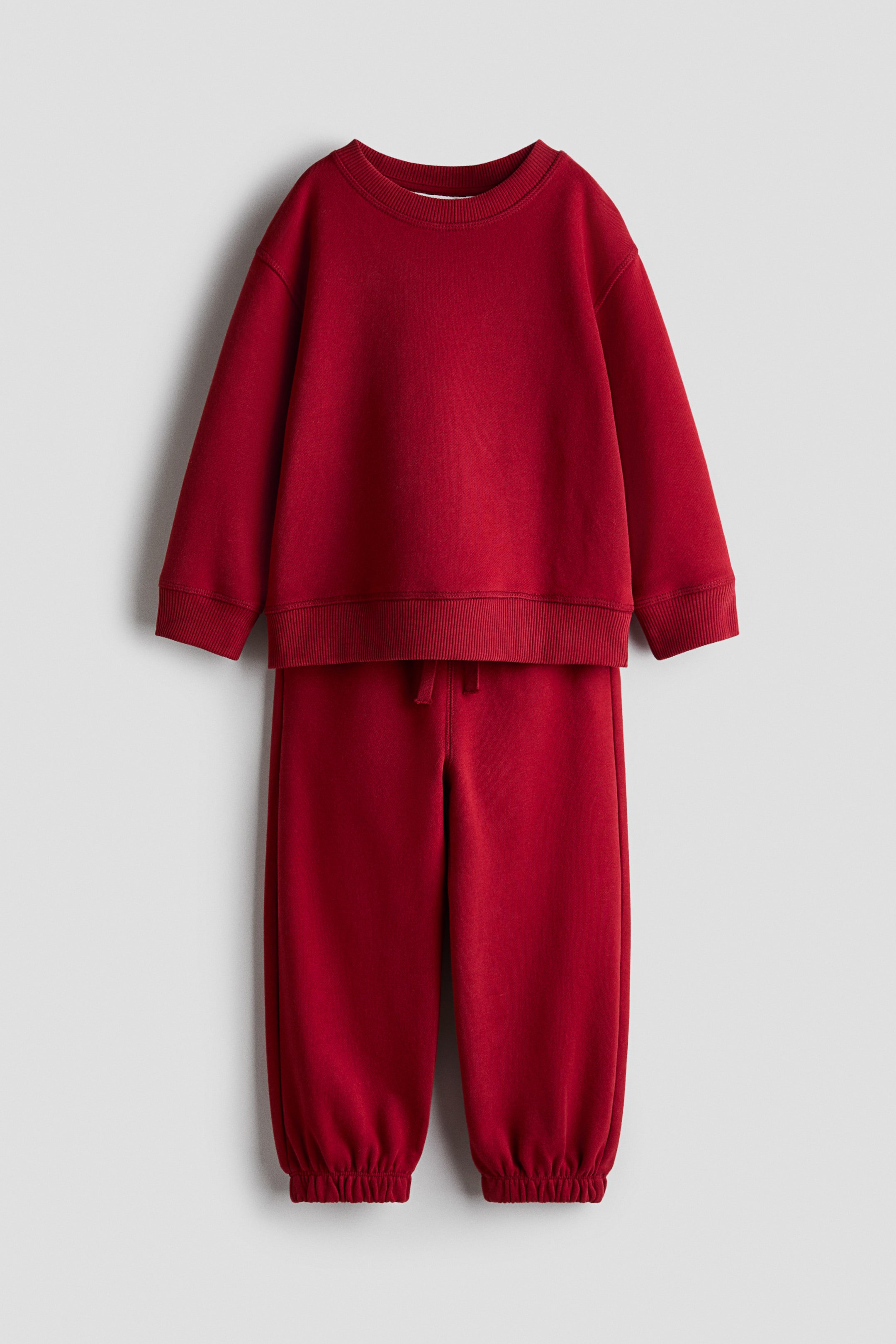 View larger image: A children’s two-piece set in deep red is shown from the front, featuring a matching crew-neck sweatshirt and joggers. The sweatshirt features dropped shoulders with wide ribbed cuffs and hem, while the joggers feature an elasticated drawstring waist and gathered elastic hems.