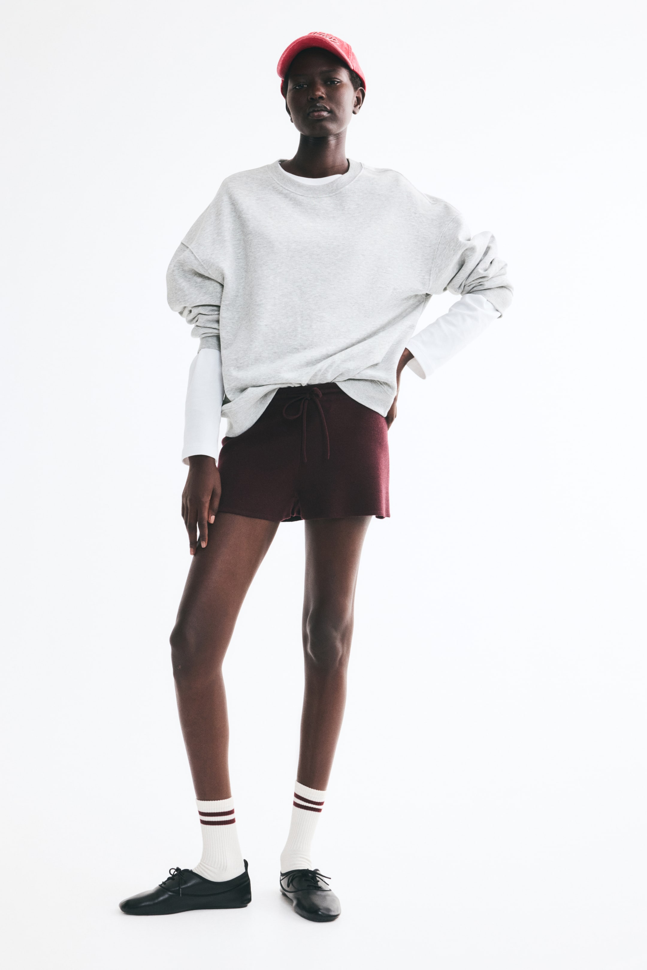 View larger image: The model is wearing an oversized light gray sweatshirt with a crew neckline, dropped shoulders, and ribbed trim. It is layered over a white long-sleeve shirt and paired with dark red drawstring shorts.