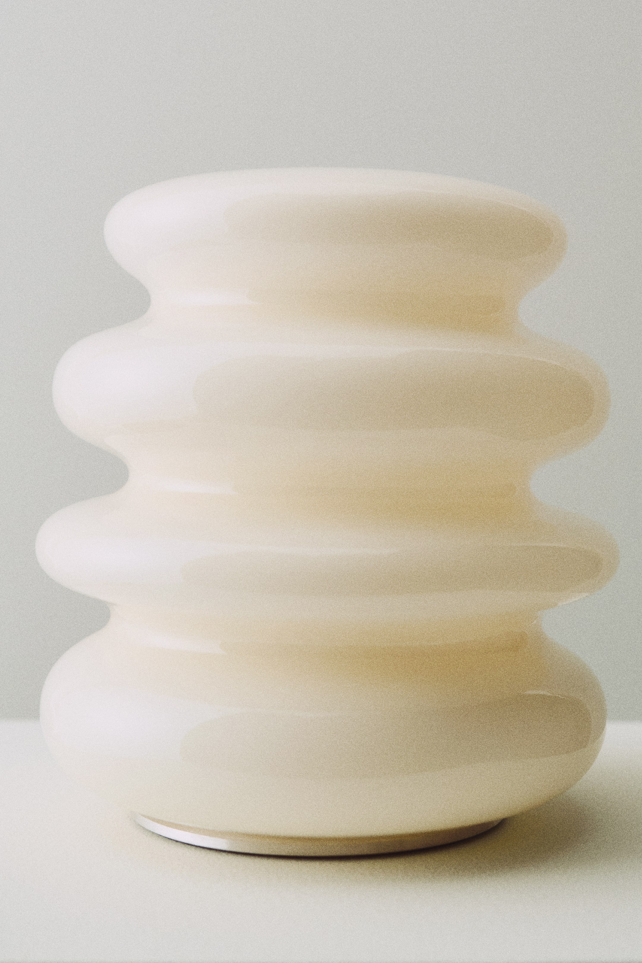 Größeres Bild ansehen: Eine wiederaufladbare Tischlampe ist frontal ausgerichtet und präsentiert einen glasfarbenen Körper in Off-White mit einem skulpturalen Design. Sie zeigt eine gestapelte, abgerundete Form mit wellenförmigen Schichten und ruht auf einem dünnen, hellfarbigen Sockel.