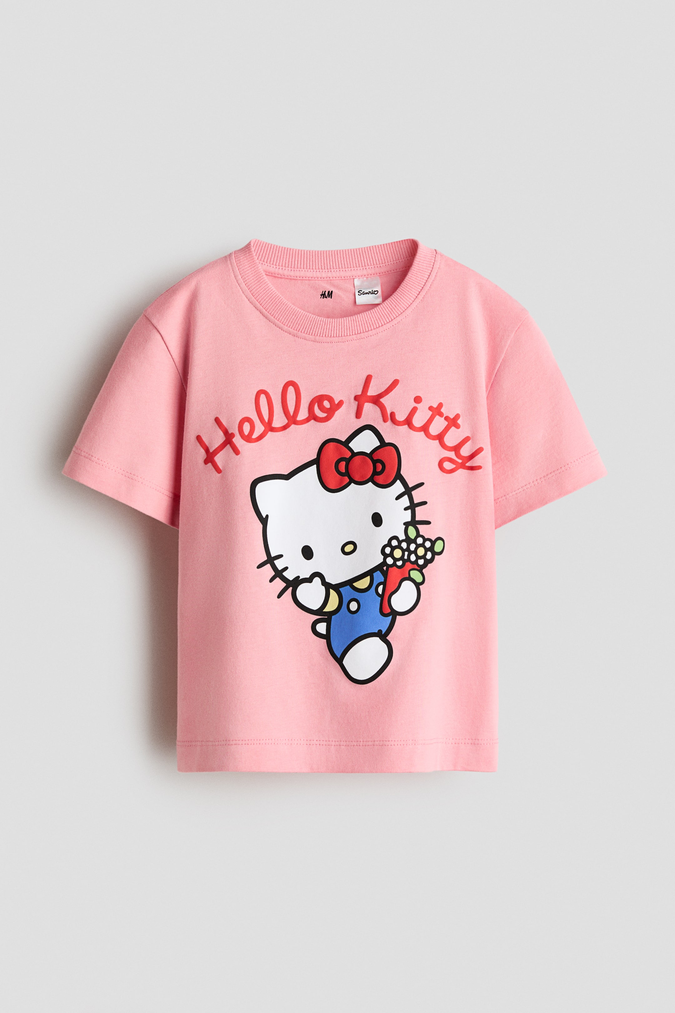 Xem ảnh lớn hơn: Áo thun màu hồng làm từ chất liệu vải cotton jersey mềm mại đang hiển thị mặt trước, với thiết kế cổ tròn có gân và tay ngắn. Mặt trước của áo có họa tiết in "Hello Kitty" bằng chữ đỏ phía trên hình ảnh Hello Kitty mặc quần yếm xanh, đeo nơ đỏ và cầm một bó hoa.