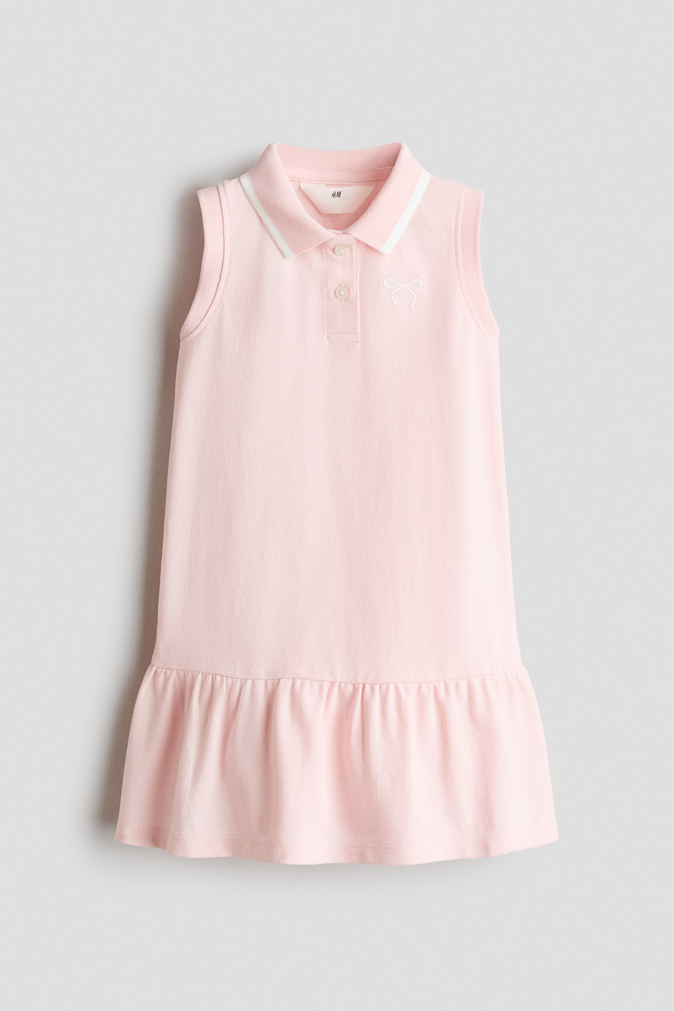 View larger image: A pink sleeveless polo dress showcasing a ribbed polo collar adorned with a white stripe and a two-button placket. A white embroidered bow detail is prominently displayed on the left chest, and the dress concludes with a ruffled flounce hem.