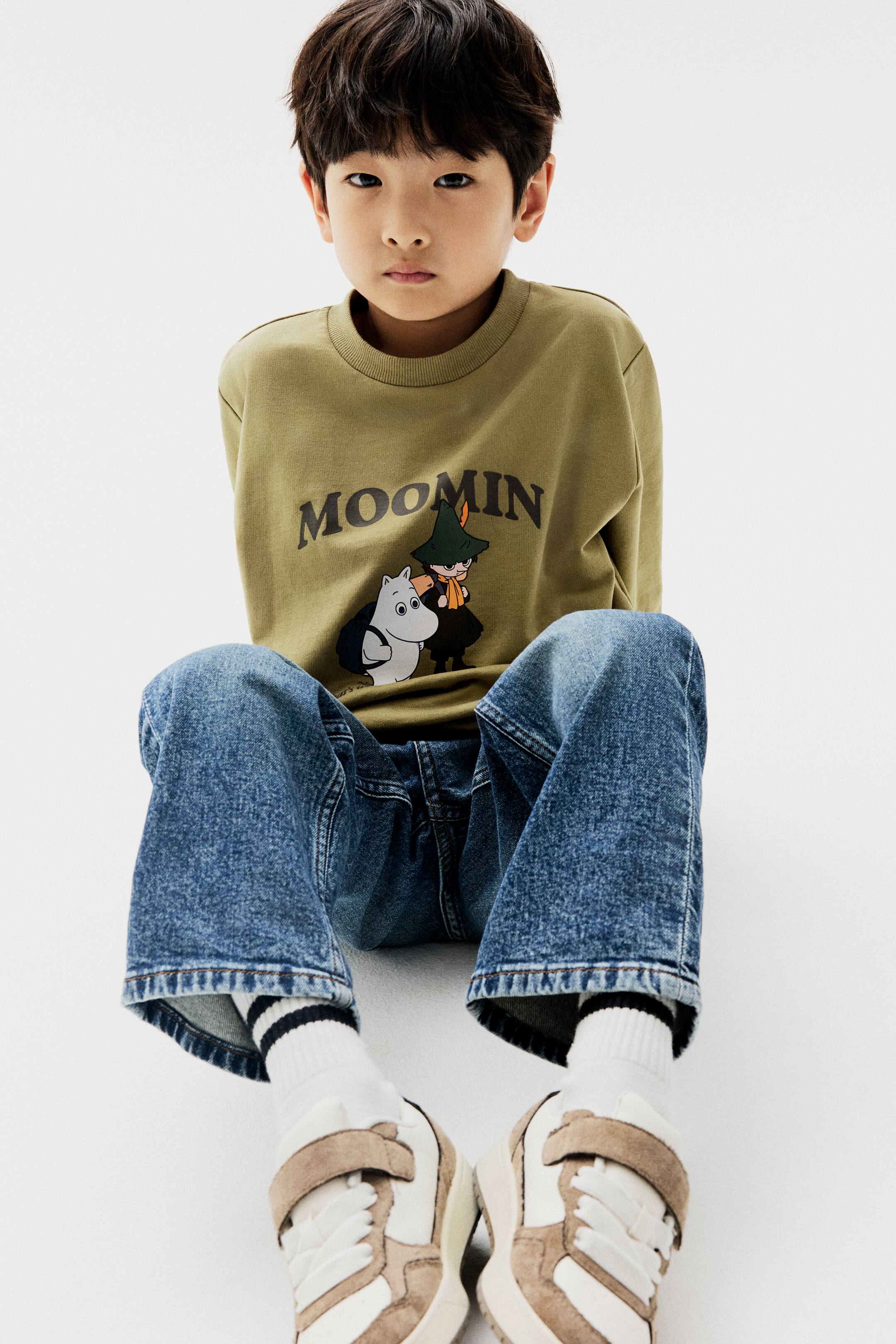 查看大圖: 模特兒正坐著穿著一件綠色輕薄衛衣,正面印有 Moomin 圖案。這件上衣採用羅紋圓領設計與落肩剪裁,並搭配藍色牛仔褲和運動鞋。