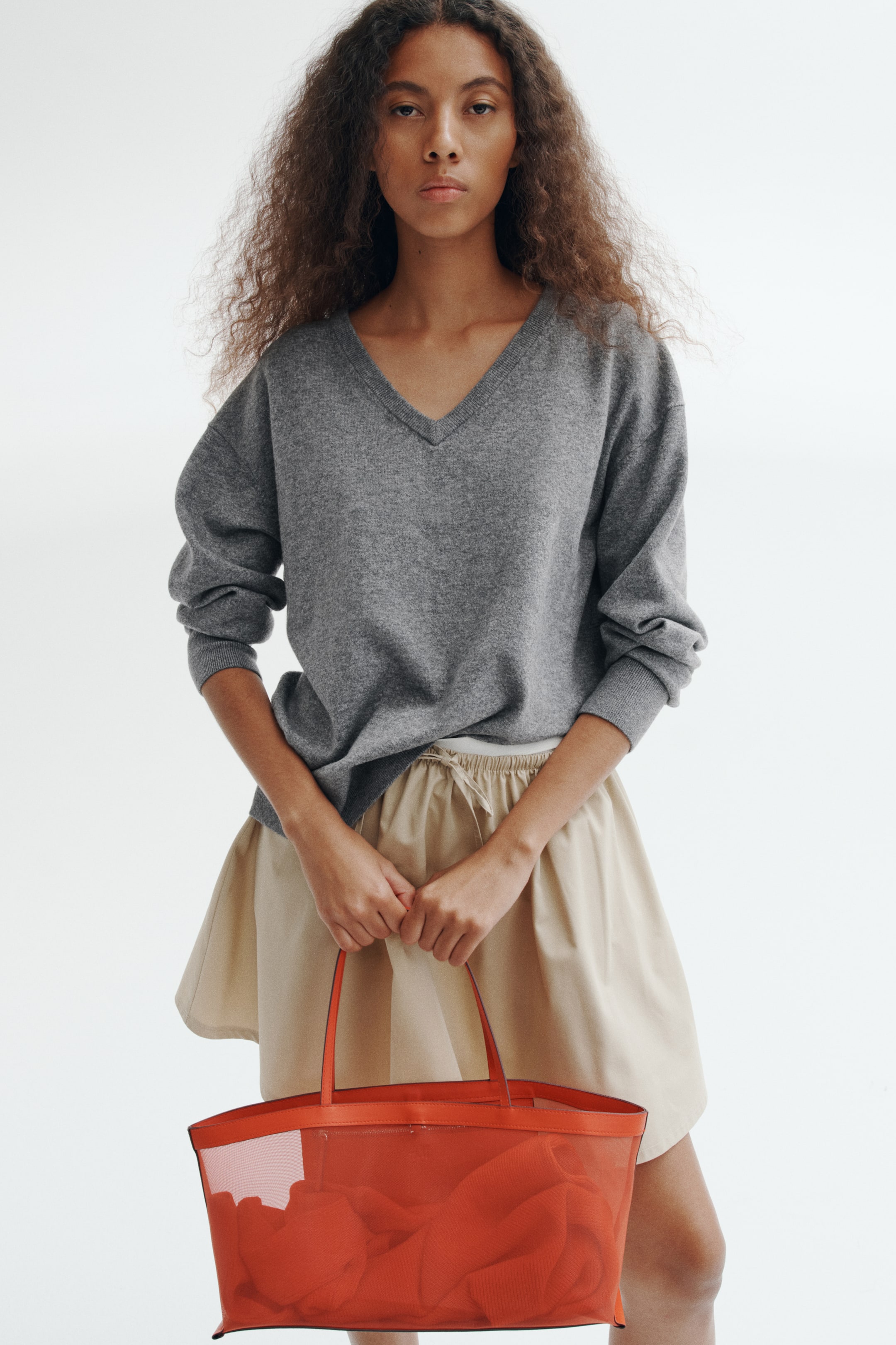 Ver imagem maior: Uma camisola oversized cinzenta de malha fina, com decote em V canelado, ombros descaídos e punhos canelados, está a ser usada pela modelo. A camisola é combinada com uma saia bege com cordão e a modelo está a segurar uma tote bag em rede em laranja-avermelhado vivo.
