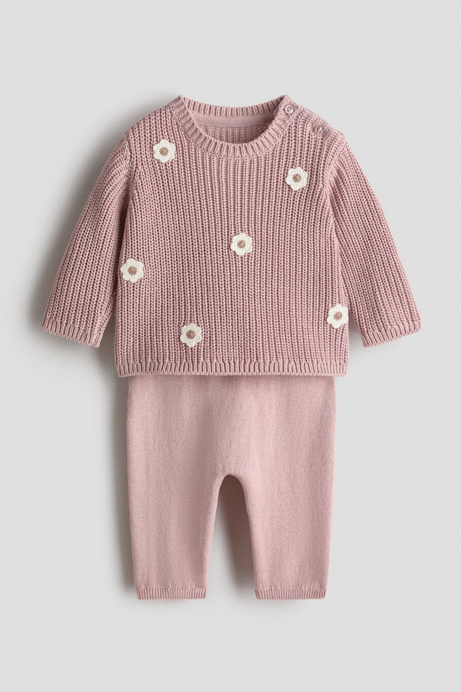 A pink knitted baby set is displaying front-facing, consisting of a ribbed knit sweater and smoother knit pants. The sweater is featuring a crew neck, two buttons on the left shoulder, long sleeves with ribbed cuffs, a ribbed hem, and white floral appliques with light brown centers. The matching pants are also featuring ribbed cuffs. - 1