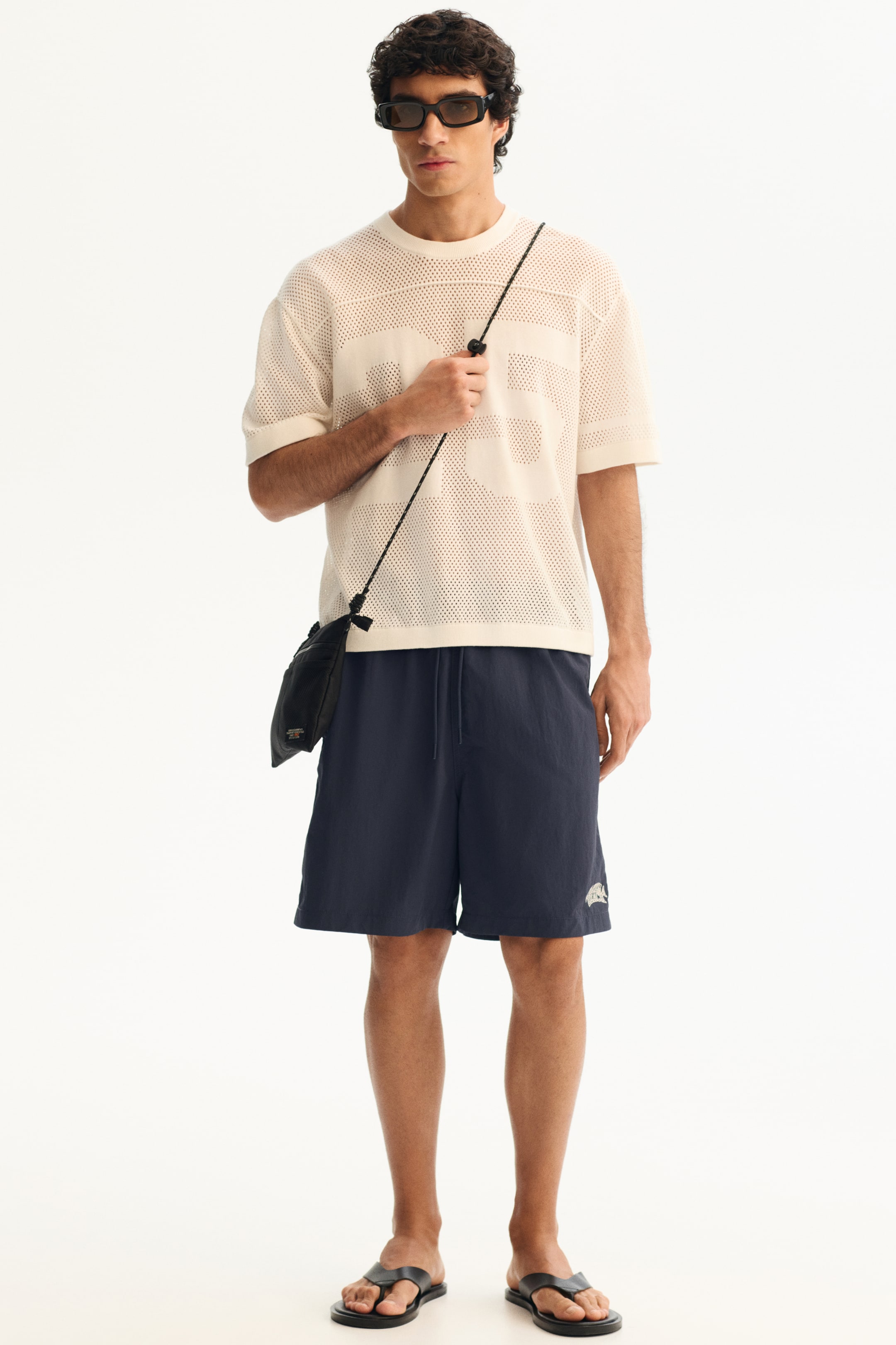 Se større billede: En cremefarvet mesh t-shirt med store '23' grafik kombineres med marineblå snøre-shorts, der falder over knæet, sammen med sorte rektangulære solbriller, en sort crossbody-taske med rebrem og sorte tåremssandaler.
