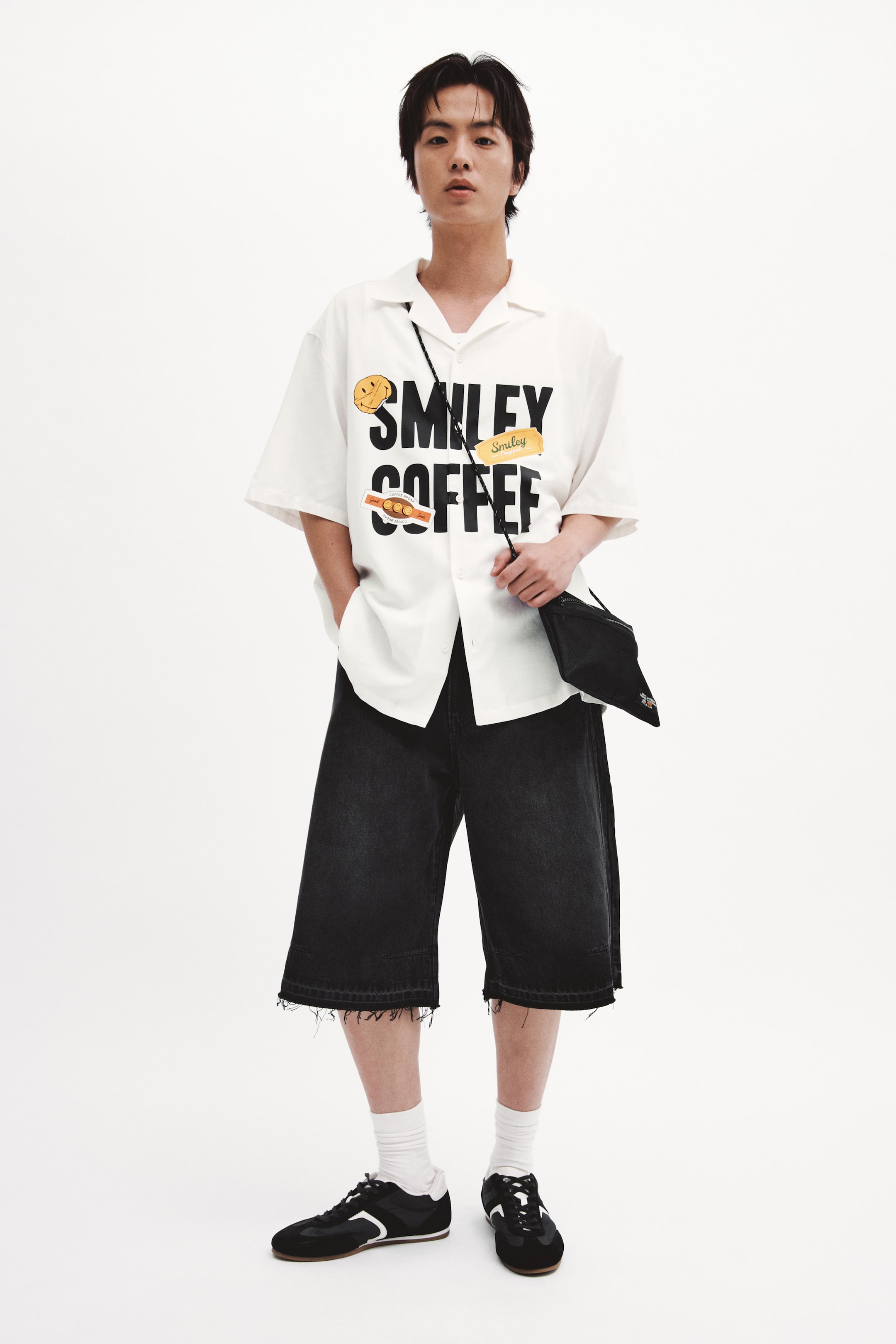 Xem ảnh lớn hơn: Áo sơ mi trắng tay ngắn có kiểu dáng rộng rãi, với hình in lớn "SMILEY COFFEE" màu đen ở phía trước. Thiết kế với cổ áo mở kiểu trại và hàng nút phía trước, áo còn có họa tiết mặt cười màu vàng, dòng chữ "Smiley" nhỏ màu vàng, cùng họa tiết hạt cà phê.