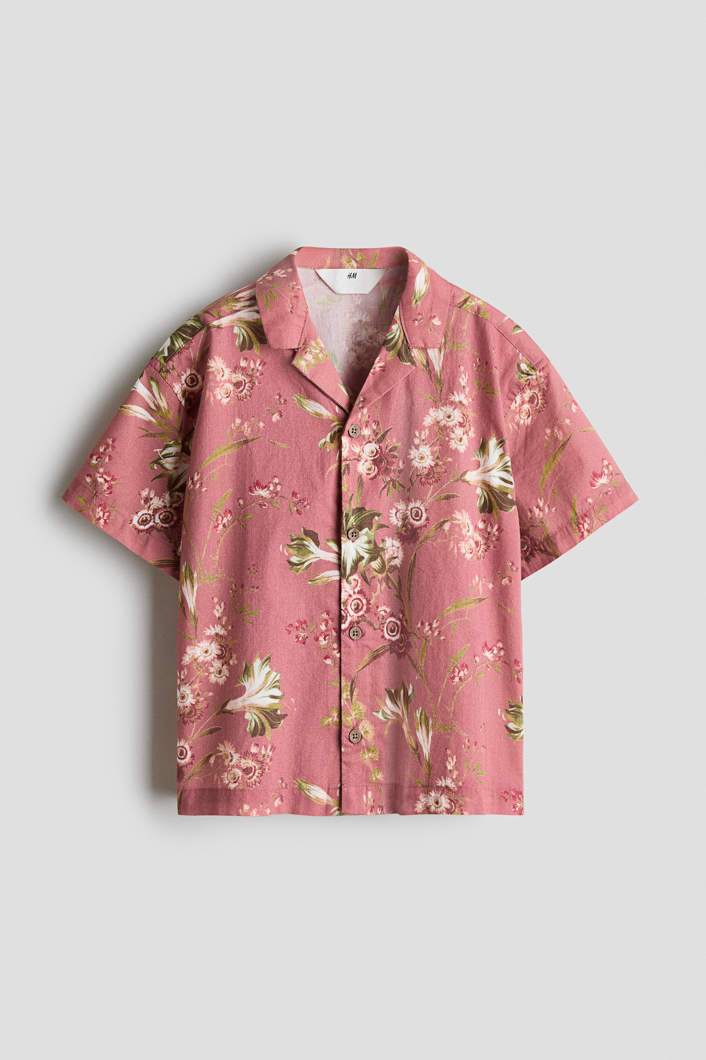 A short-sleeve shirt in dusty pink cotton weave with an all-over floral print is worn facing front. The shirt features a resort collar, a button-down front, and gently dropped shoulders, draping loosely over brown trousers.