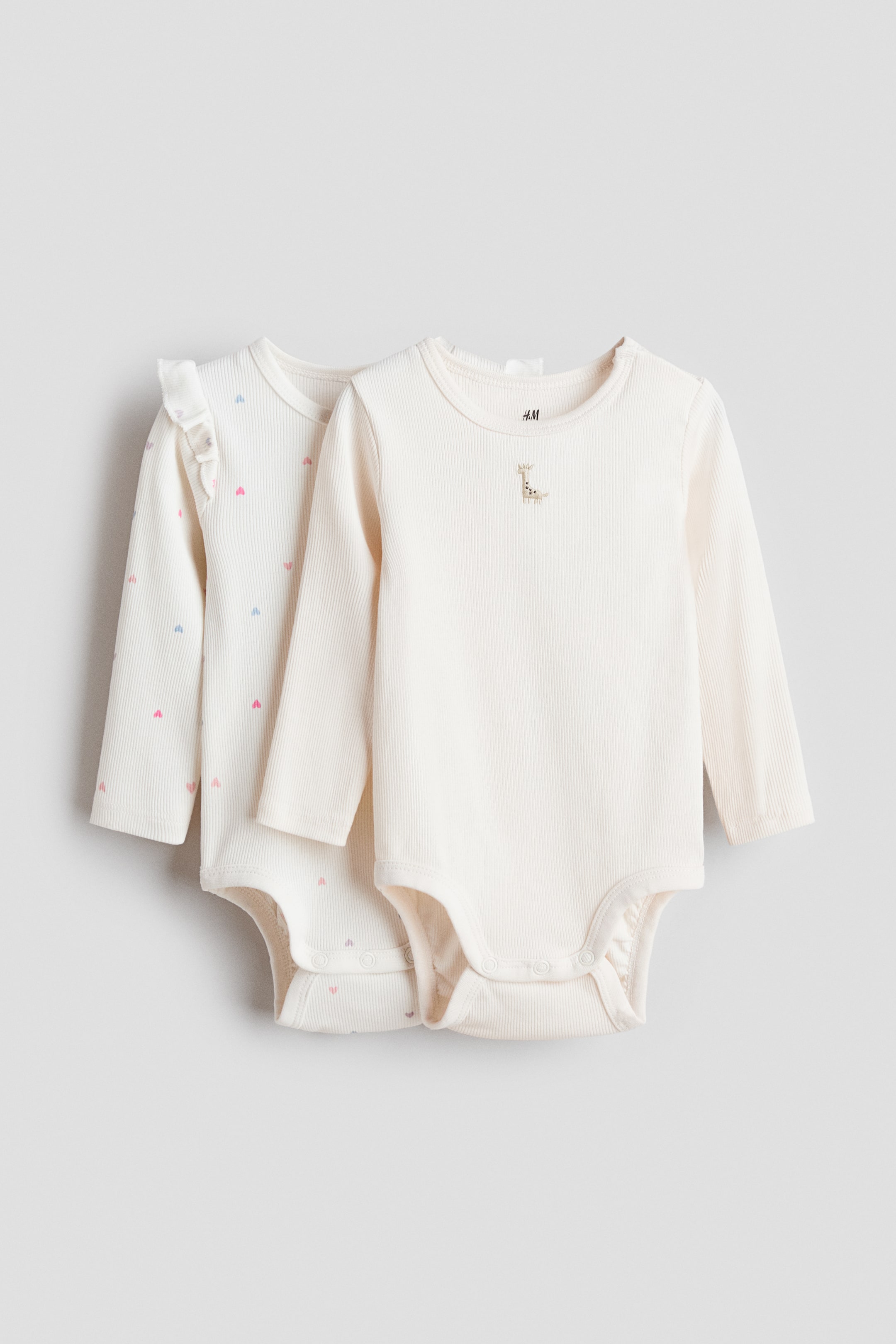 Größeres Bild anzeigen: Zwei gerippte Baby-Bodys präsentieren ihre Vorderseiten. Der vordere Body ist ein schlichtes, cremefarbenes Langarm-Design mit einer kleinen gestickten Giraffe auf der linken Brust, während der Body dahinter ein cremefarbenes Langarm-Design mit kleinen rosa und blauen herzförmigen Punkten und Rüschenverzierungen an den Schultern ist. Beide Bodys weisen Druckknopfverschlüsse im Schritt auf.