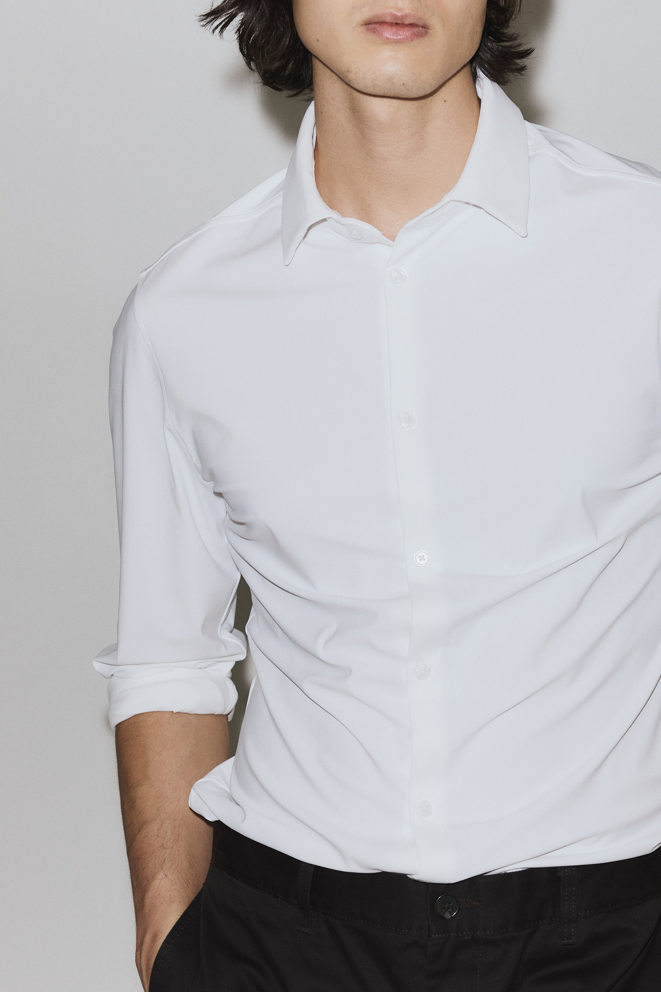 Ampliar la imagen: Una camisa blanca de manga larga presenta un cuello clásico tipo spread y una botonadura frontal con botones blancos. Las mangas están remangadas hasta los antebrazos y la camisa está metida dentro de pantalones oscuros, mostrando un aspecto liso y entallado.