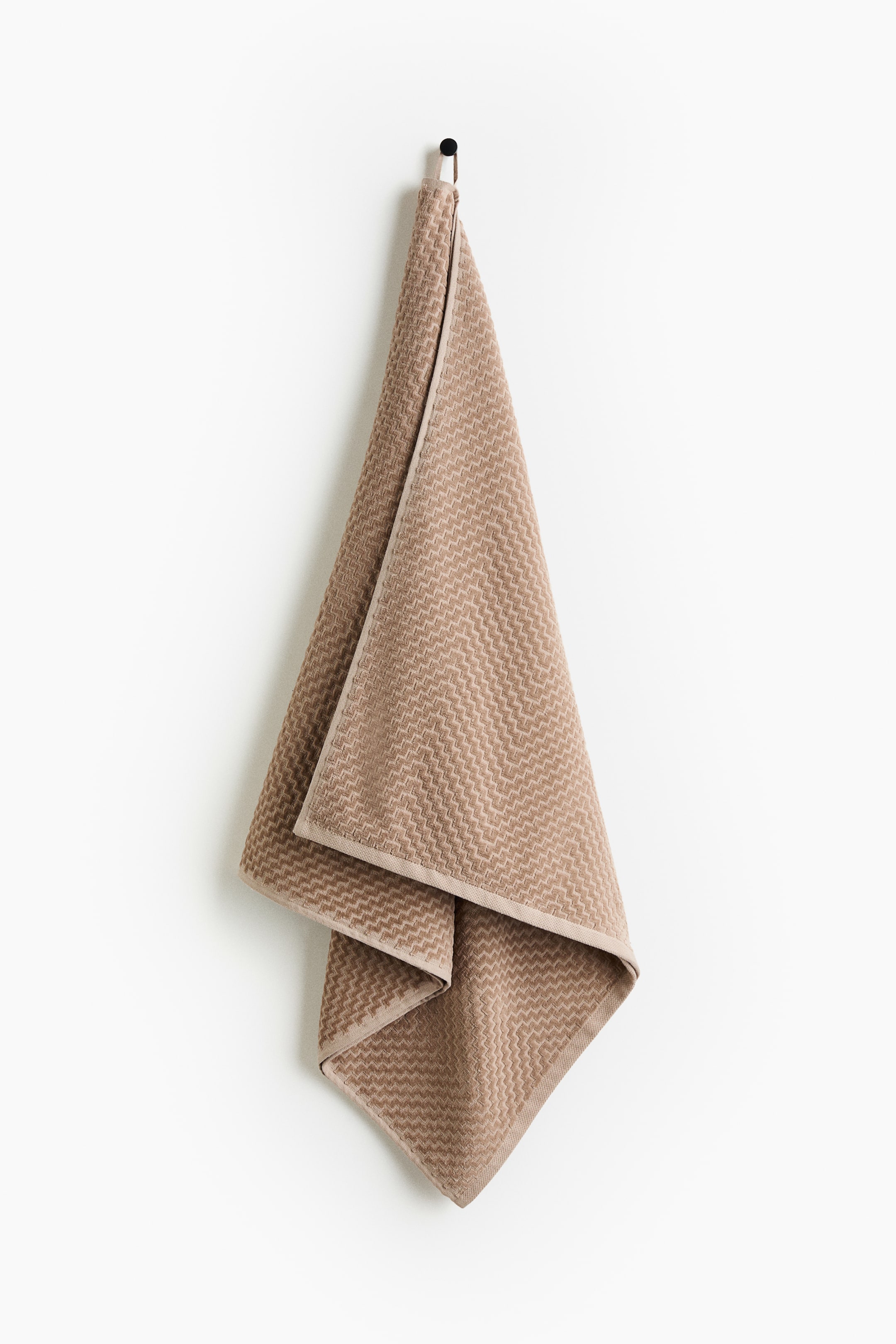 View larger image: A light brown bath towel, showcasing a textured zigzag pattern, is suspended from a dark hook by its integrated loop. The towel is folded, displaying its layers and delicate edge trim.