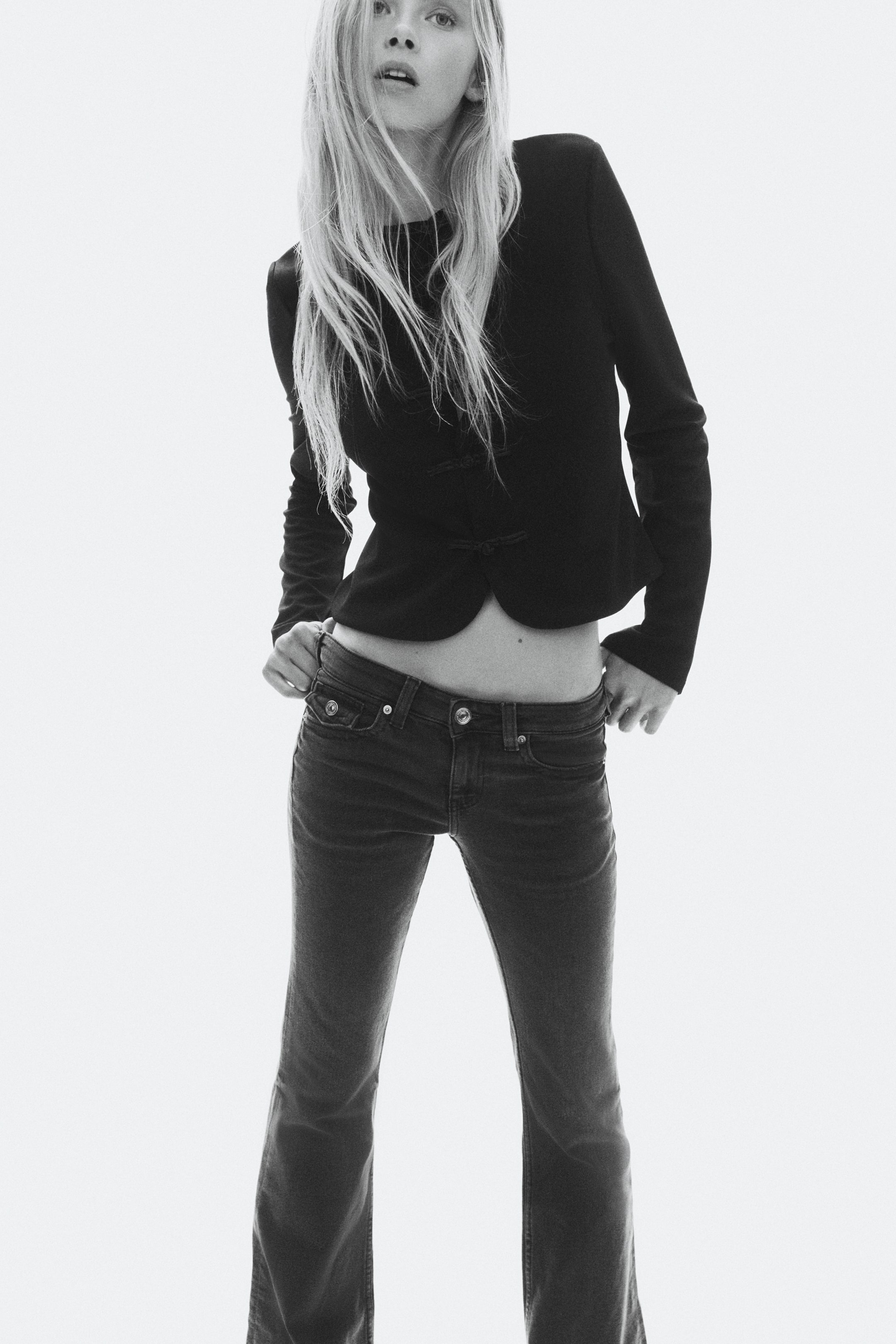 View larger image: The model is wearing dark denim jeans with a low waist, slim-fit thighs, and a flared cut from the knees, holding the waistband with both hands. The jeans are styled with a long-sleeved black jacket, cropped to reveal the midriff.