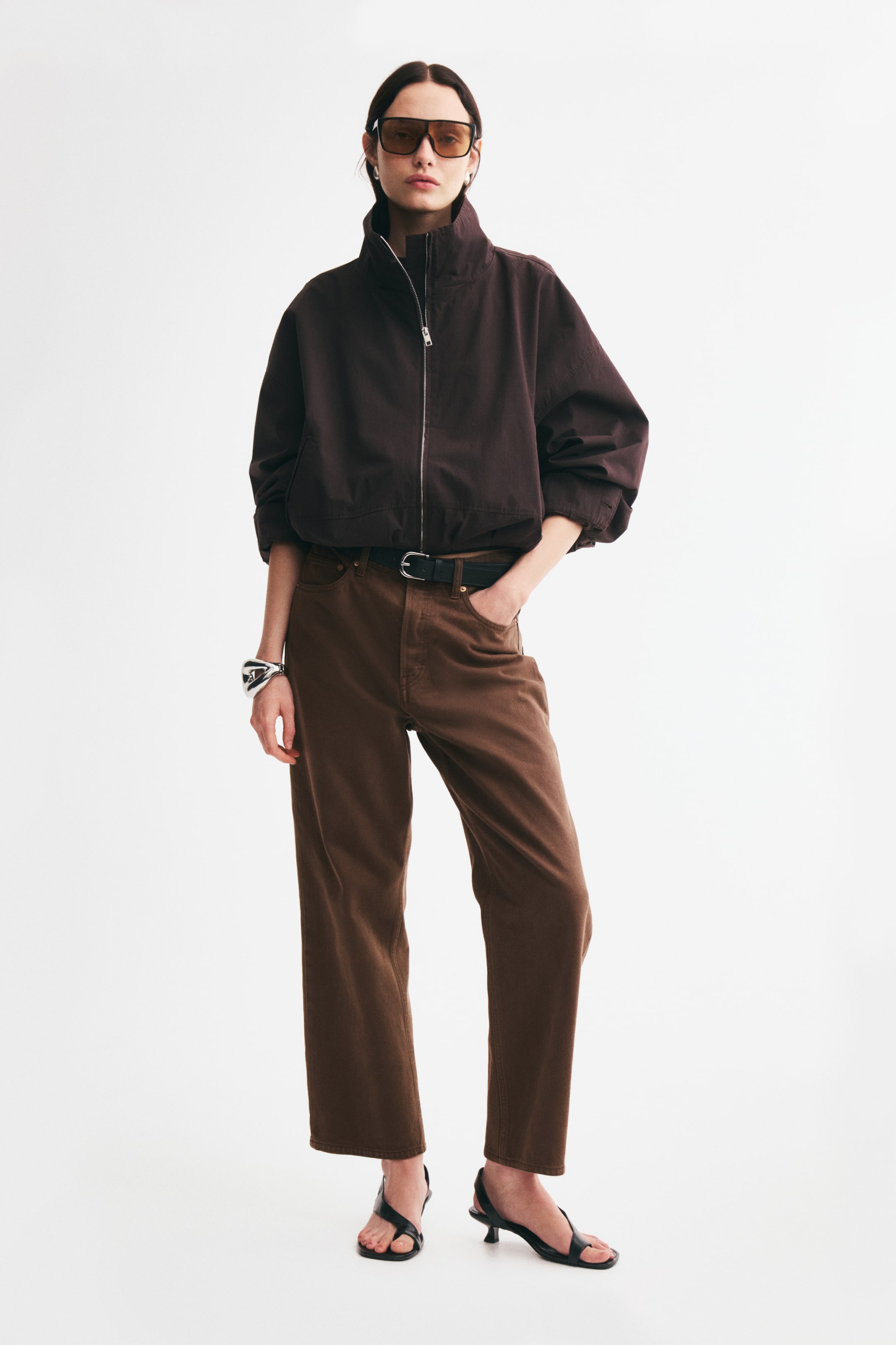 View larger image: The model is wearing a dark brown high-neck zip-up jacket and brown straight-leg jeans. The jacket features a silver front zipper and gathered sleeves, while the jeans are styled with a black belt, with one hand resting in the front pocket.