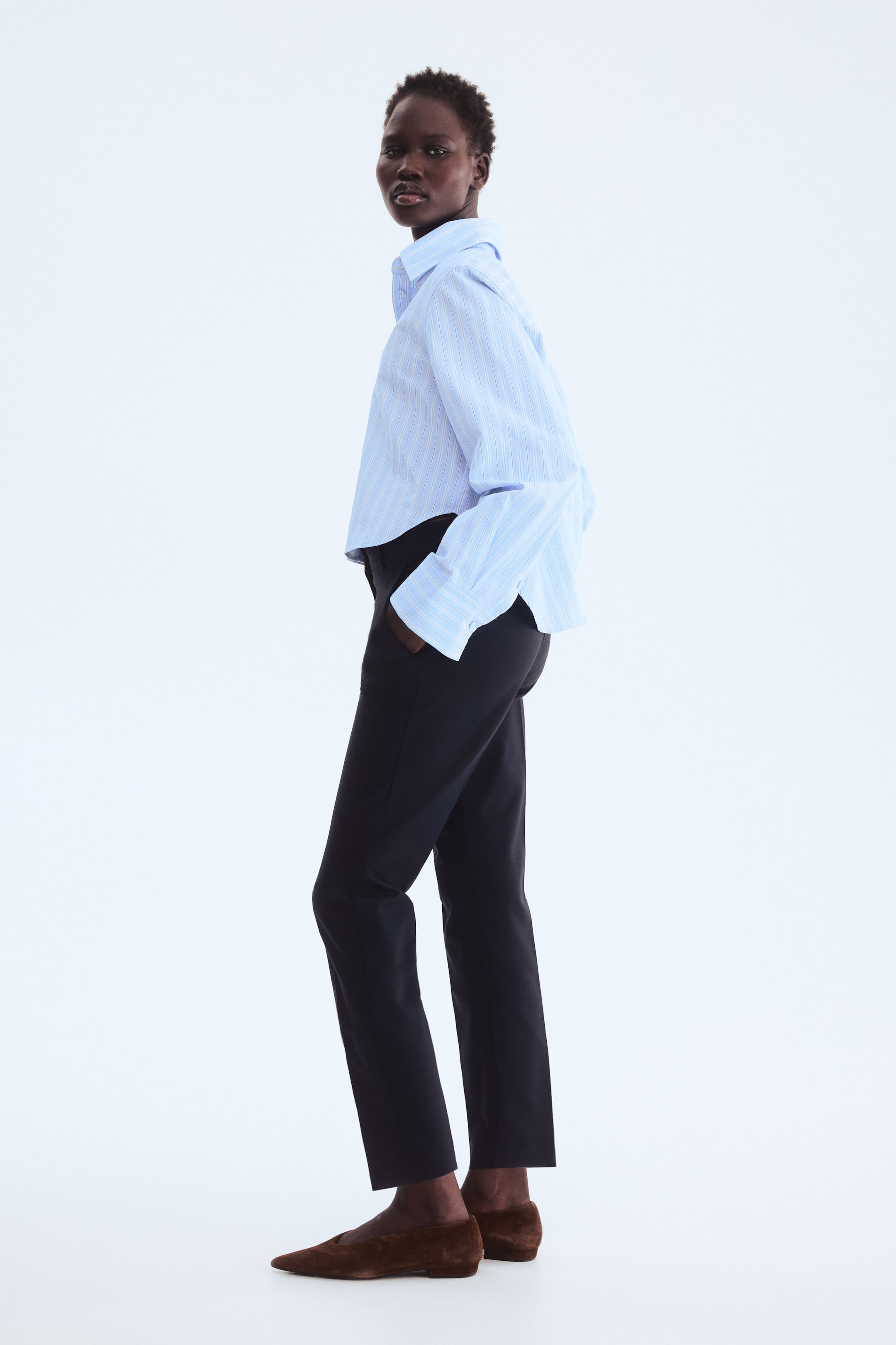 Grotere afbeelding bekijken: Een lichtblauw en wit gestreept cropped shirt wordt gedragen, met een kraag en lange mouwen met knoopmanchetten, en heeft een ontspannen pasvorm. Het is gecombineerd met donker, slim-fit enkel-lange broeken en bruine suède puntige flats.
