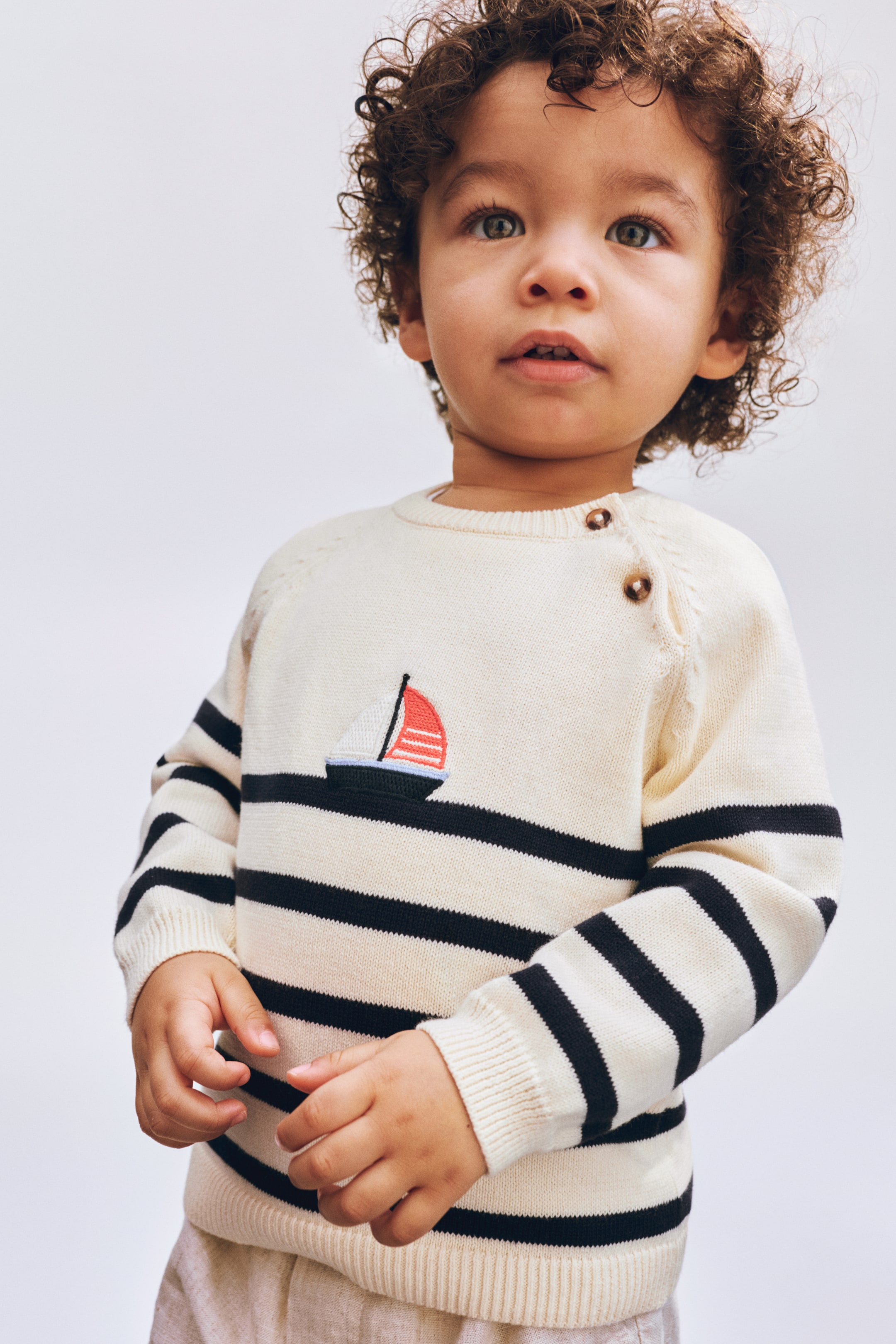 View larger image: A cream-coloured jumper, featuring black horizontal stripes and an embroidered sailboat with a black hull and red and white sail on its front, is displaying two brown buttons on the left shoulder. The jumper is also showing ribbed cuffs and a ribbed hem.