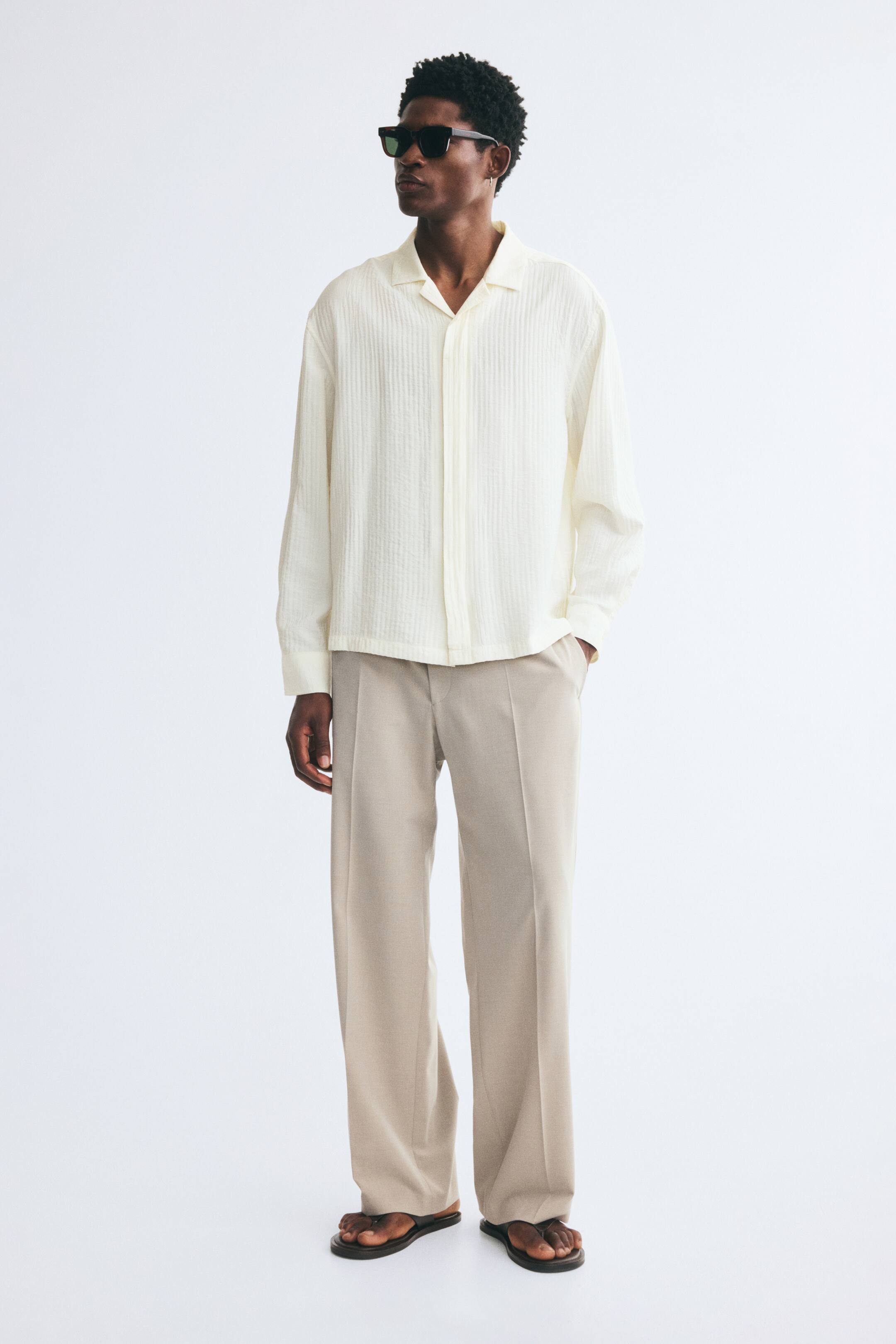View larger image: An off-white shirt in an airy, textured weave is worn by the model, featuring a relaxed fit, a resort collar, and long sleeves with button cuffs. The shirt is styled untucked with loose-fitting beige trousers and dark sandals.