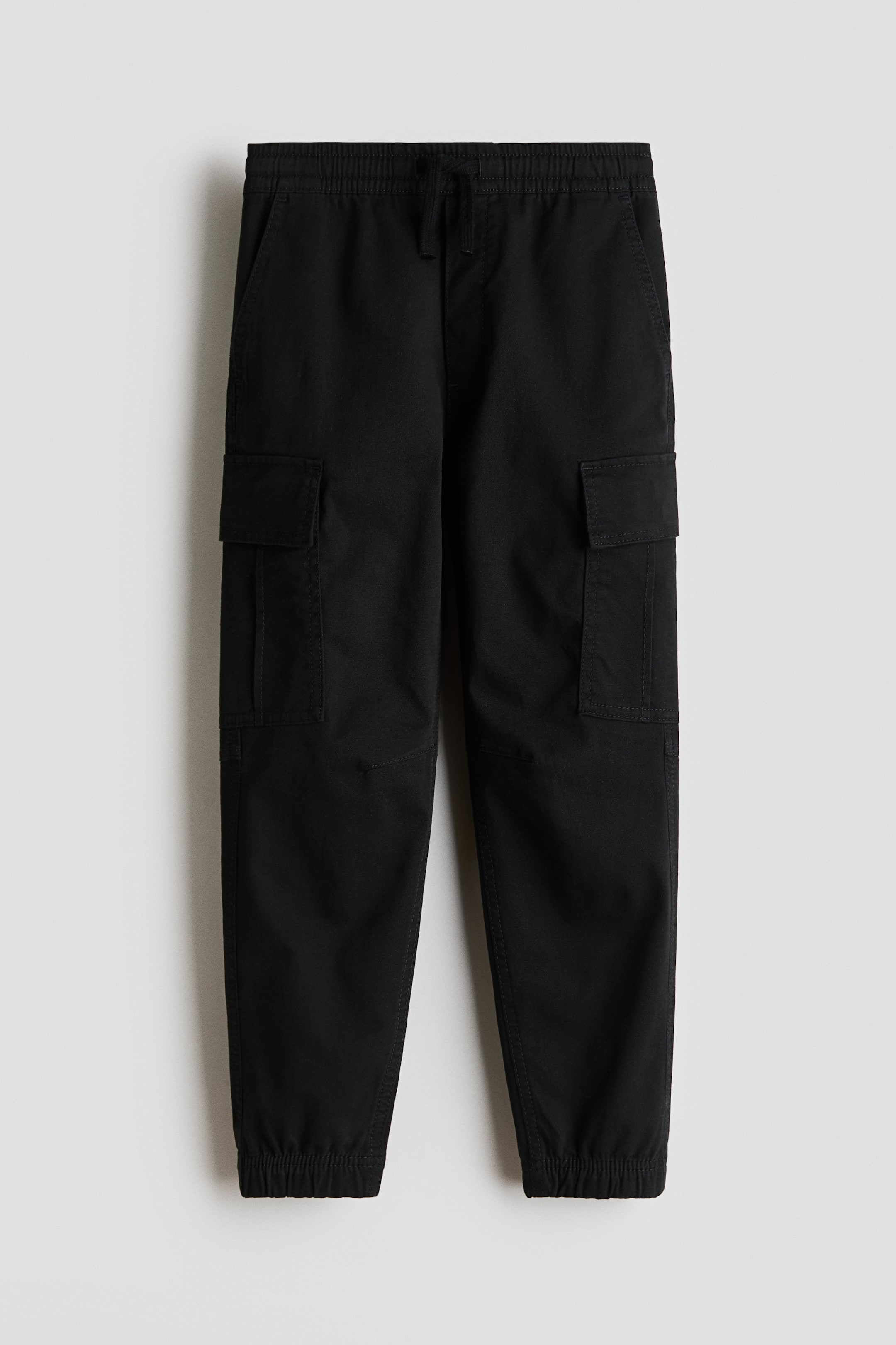 Voir une image plus grande: Pantalons cargo noirs avec taille élastiquée et cordon de serrage, deux poches avant en biais et deux poches cargo à rabat sur les côtés, complétés par des poignets élastiqués aux chevilles.