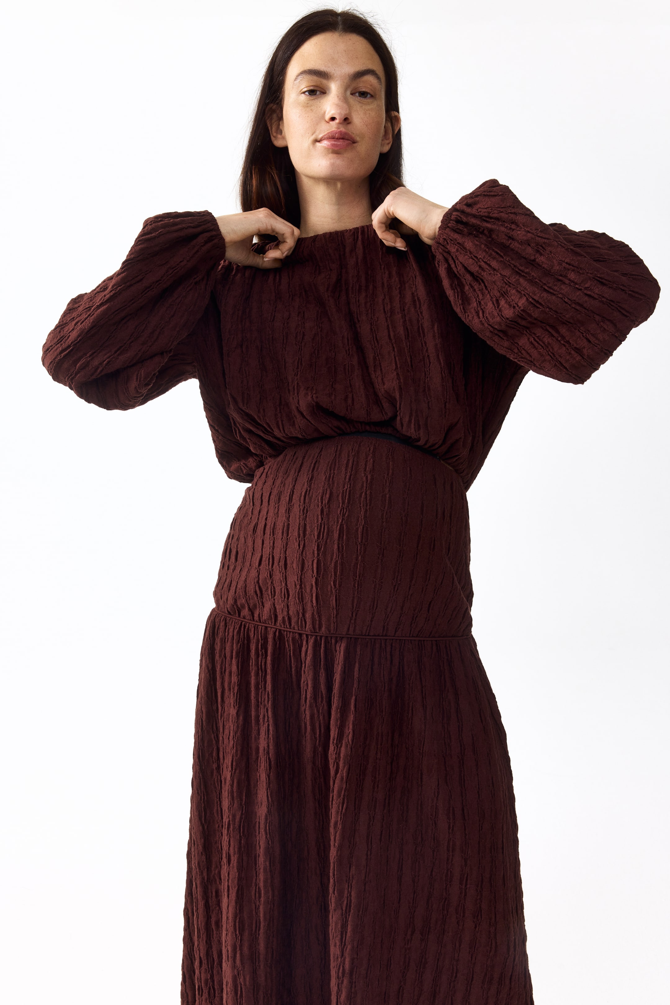 View larger image: "A deep mahogany dress is being worn, showcasing a crinkled texture across its flowy bodice, long billowy sleeves, elasticized waist, and a gathered skirt, with hands adjusting the high neckline."