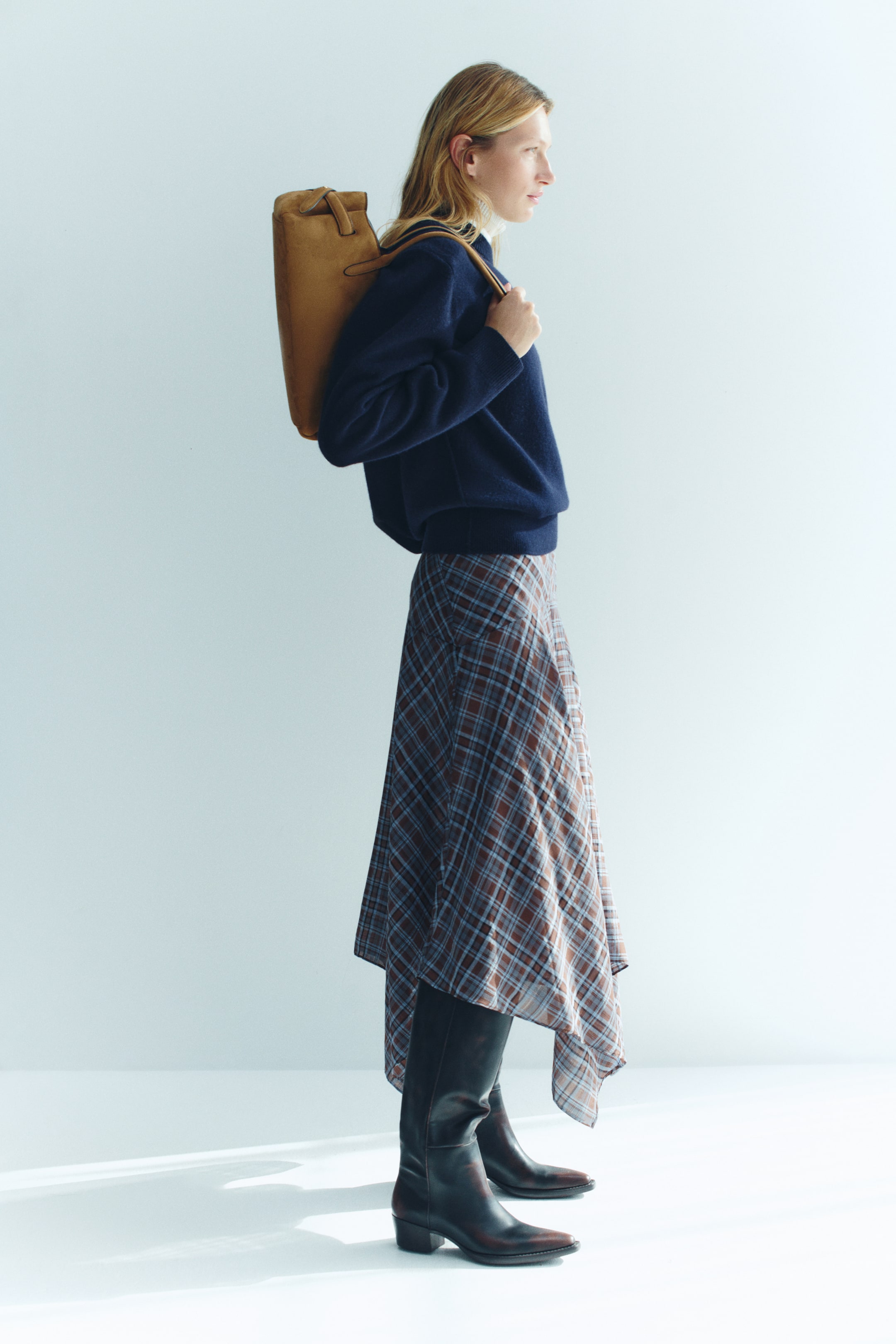 View larger image: A calf-length skirt in blue and brown plaid woven fabric, featuring an asymmetrical handkerchief hem, is worn by the model. The skirt is styled with a navy blue knit sweater and tall black leather boots.