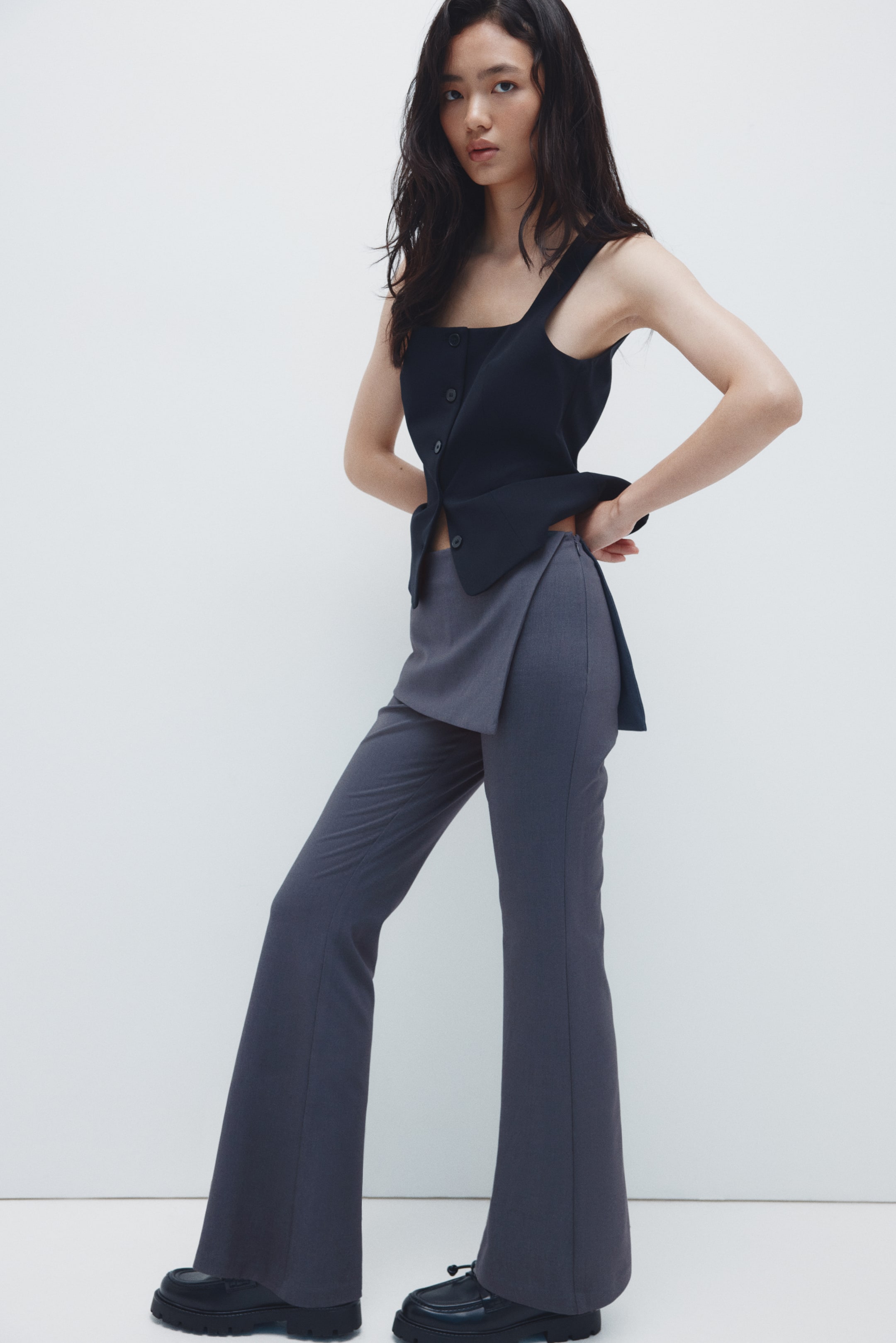 View larger image: A dark navy sleeveless top is showcasing a square neckline and an asymmetrical button-front closure, with its asymmetrical hem draping over dark gray wide-leg pants. The pants are featuring a unique, overlapping front panel extending from the waist and are flaring out from the knee.