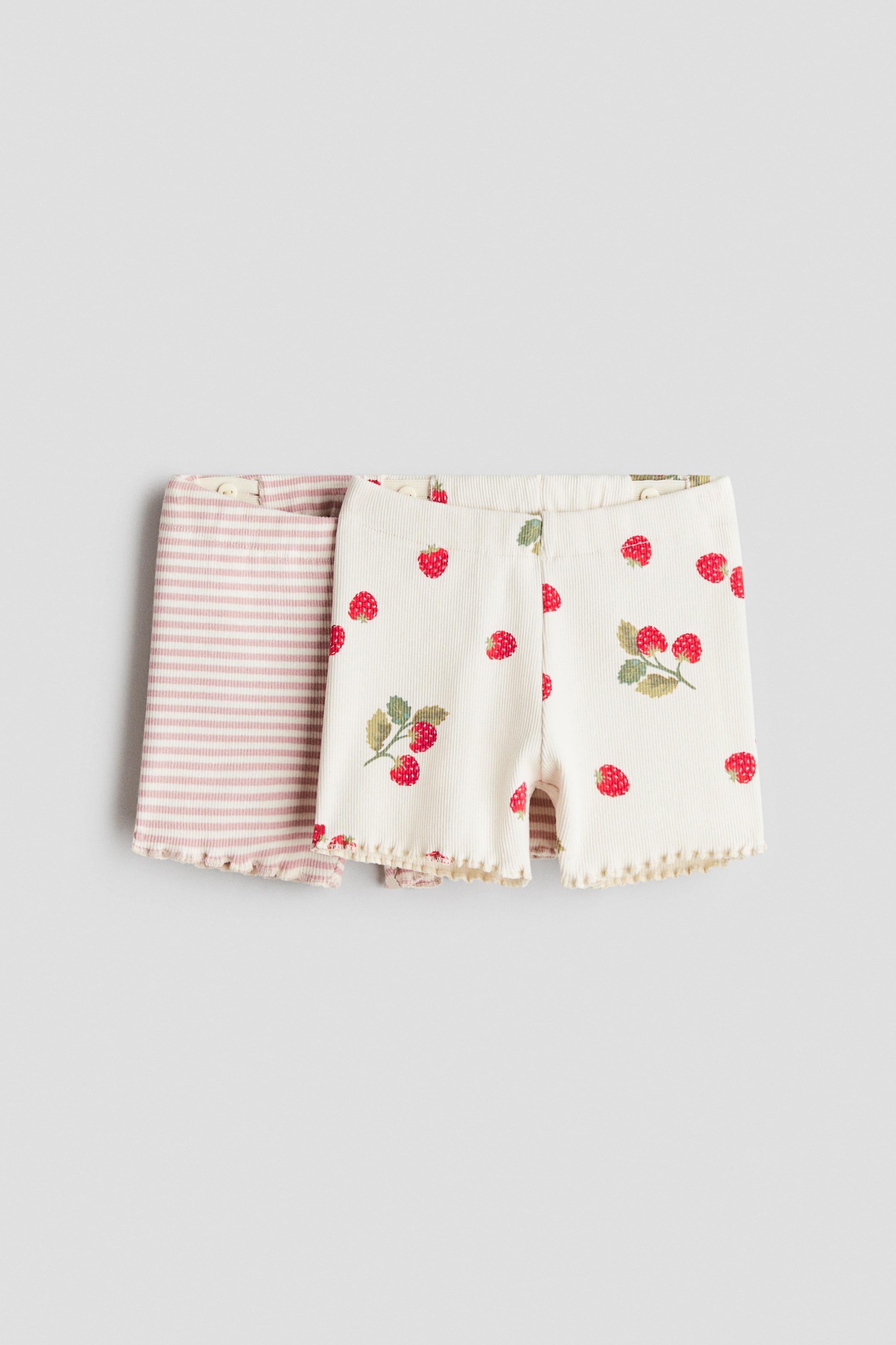 View larger image: Two pairs of H&M Adorables cycling shorts are shown from the front, both featuring a ribbed jersey texture. One pair is light pink with white horizontal stripes, and the other is cream with red strawberry patterns, both featuring an elastic waistband and wavy overlocked trims at the hems.