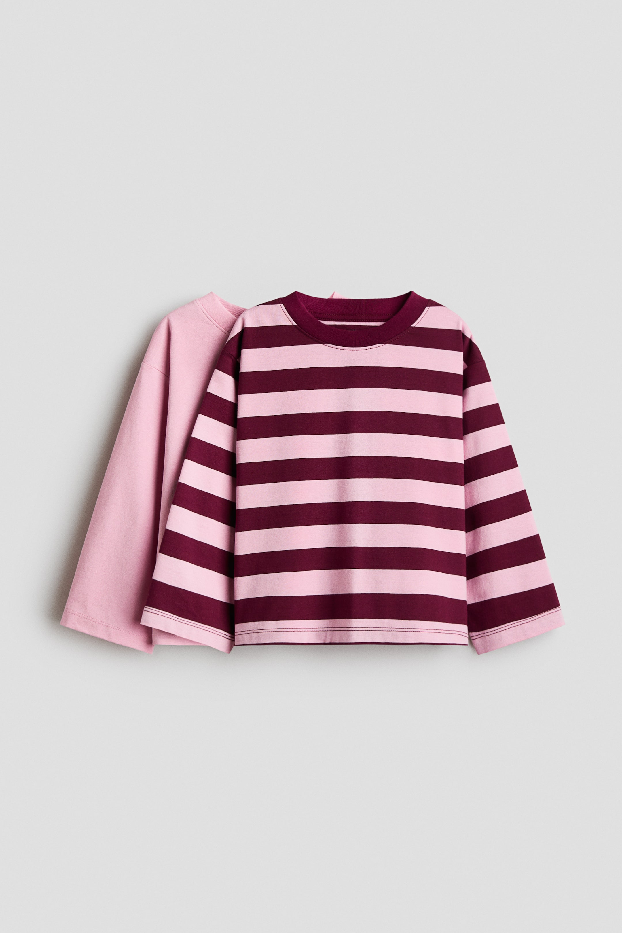 View larger image: Two relaxed-fit, long-sleeved T-shirts are shown from the front, with the front shirt featuring horizontal stripes in light pink and dark burgundy, a burgundy ribbed crew neck, and dropped shoulders. The rear T-shirt, partially visible behind the striped one, is solid light pink.