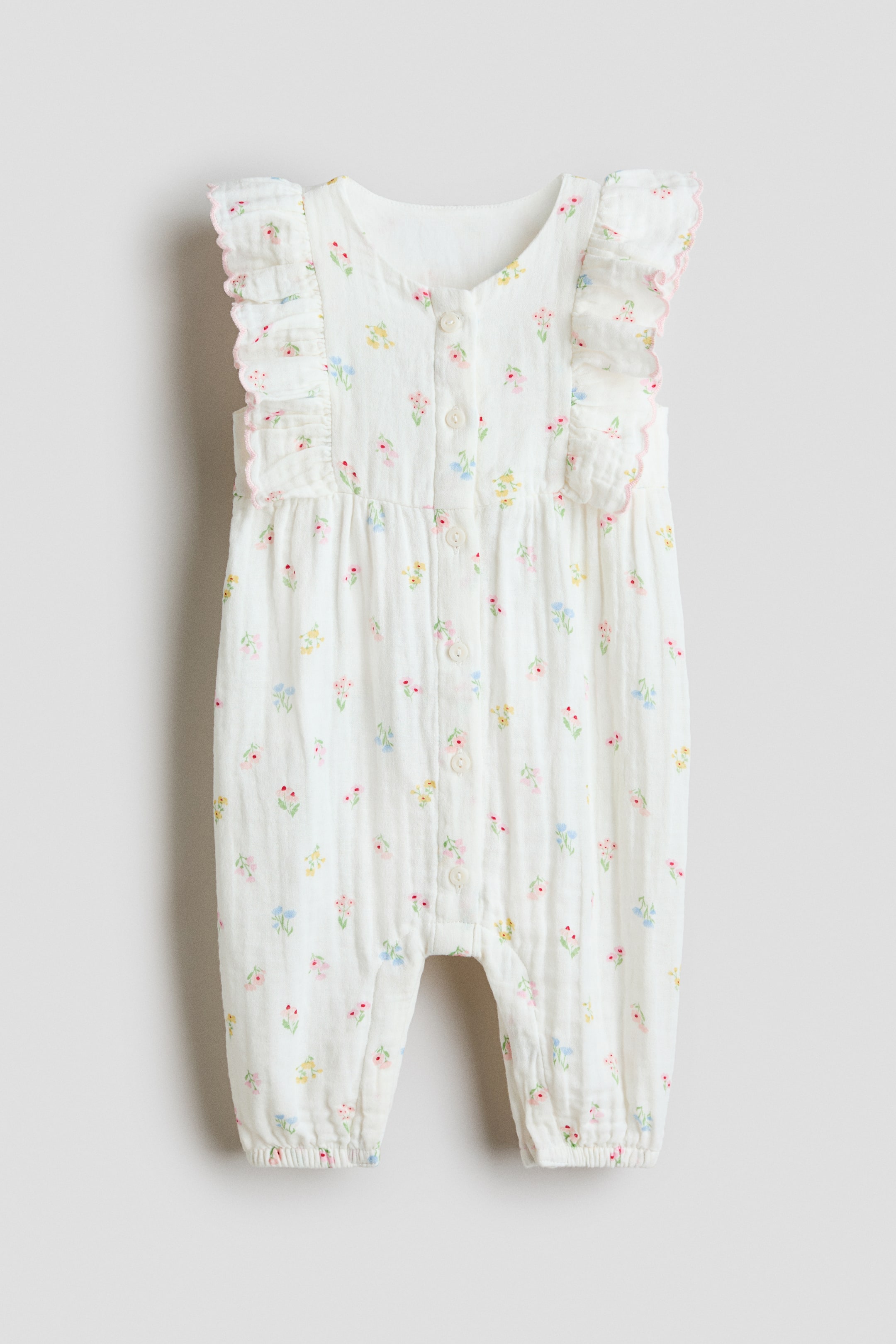 View larger image: A frilled muslin romper suit is shown from the front, showcasing a white fabric adorned with a scattered small floral print in pink, yellow, and blue. It features frilled shoulder straps accented with a pink picot trim, a front button placket, and gathered leg cuffs.