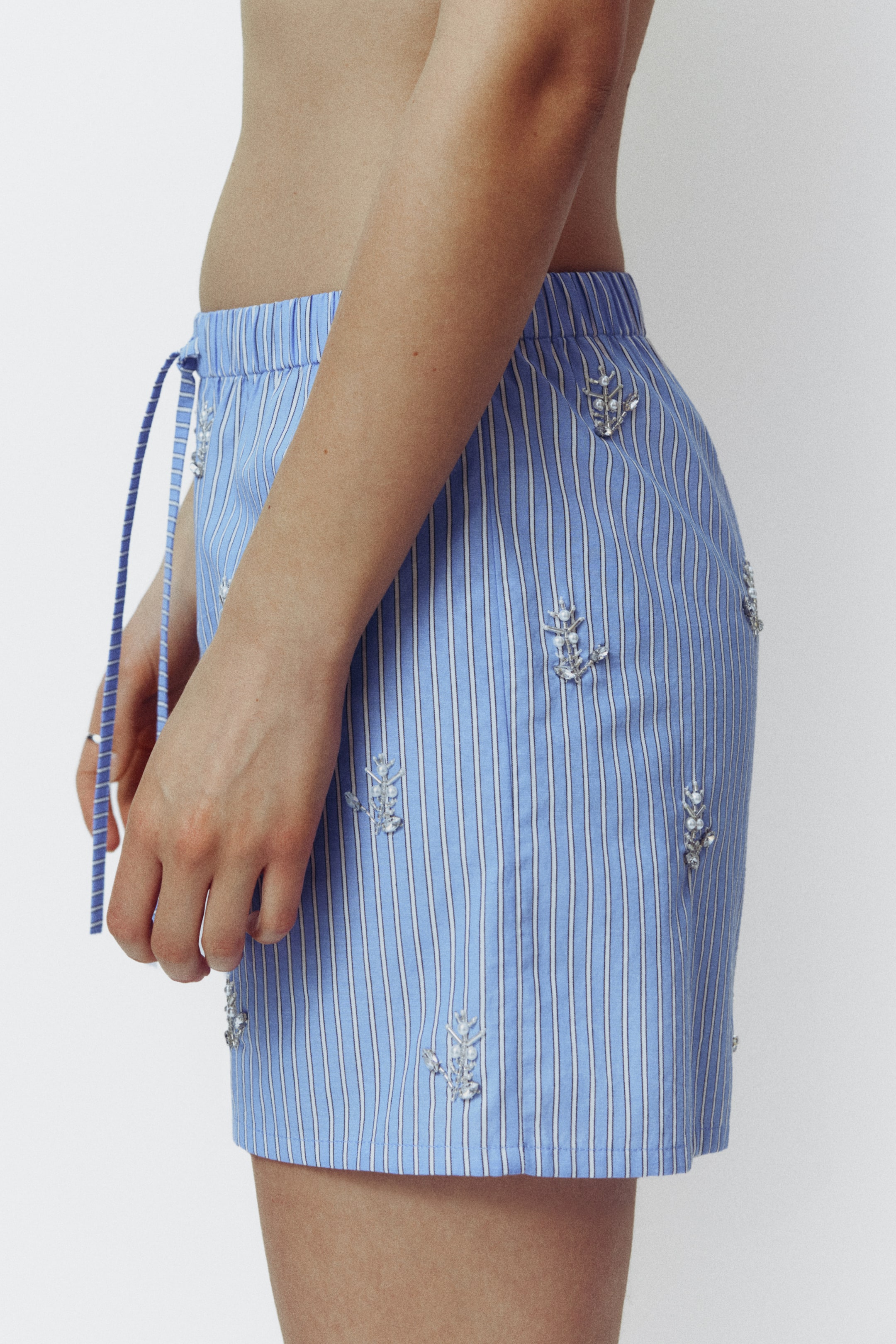 Voir une image plus grande: Les shorts sont bleus avec des rayures verticales blanches, dotés d'une taille élastique avec cordon de serrage et de délicates ornements perlés argentés. Ils sont présentés de côté, mettant en valeur la coupe et le design.
