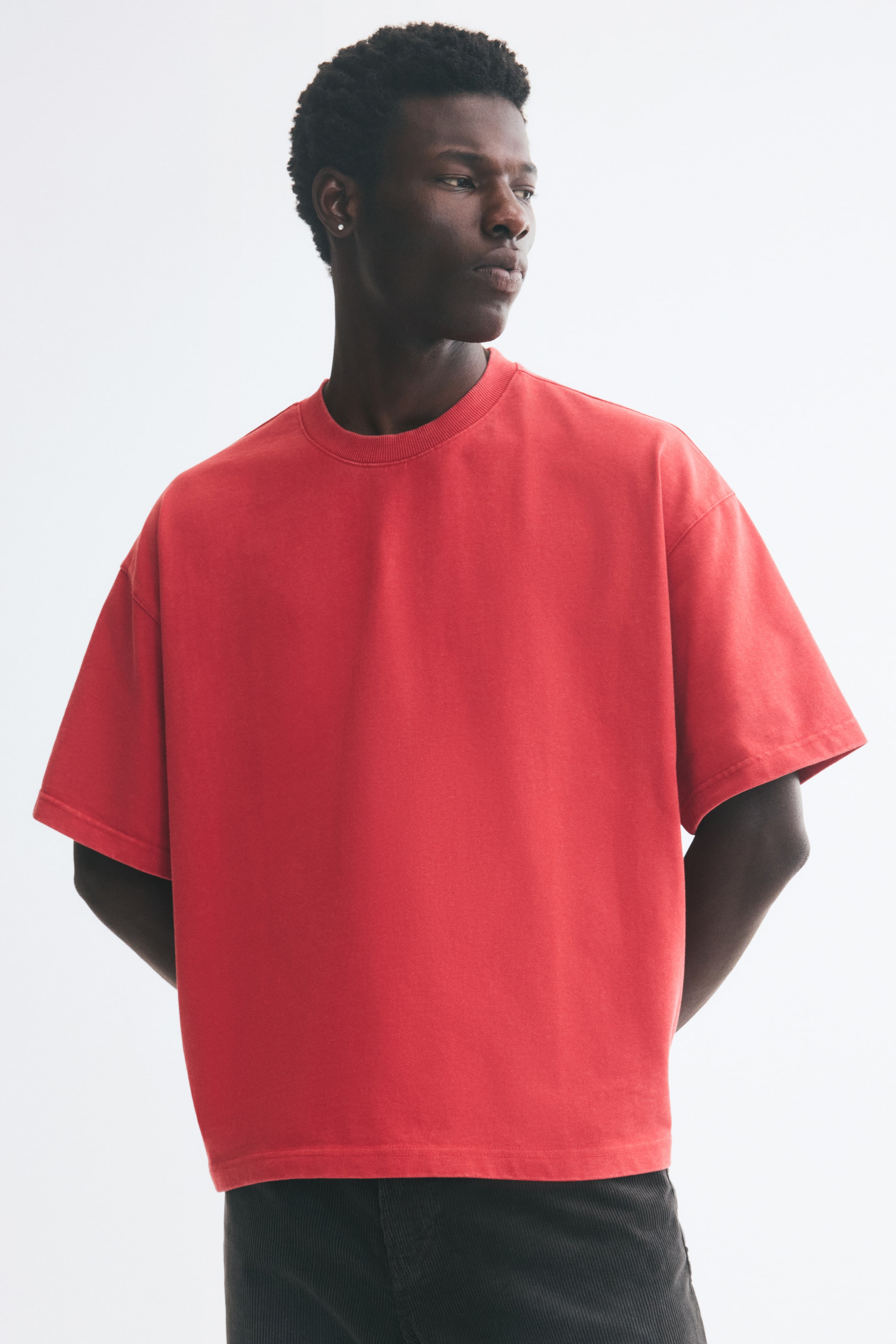 View larger image: A red, boxy-style T-shirt is being worn by the model, featuring a ribbed crew neckline, dropped shoulders, and wide short sleeves. The oversized tee is paired with dark trousers as the model stands with hands positioned behind their back.