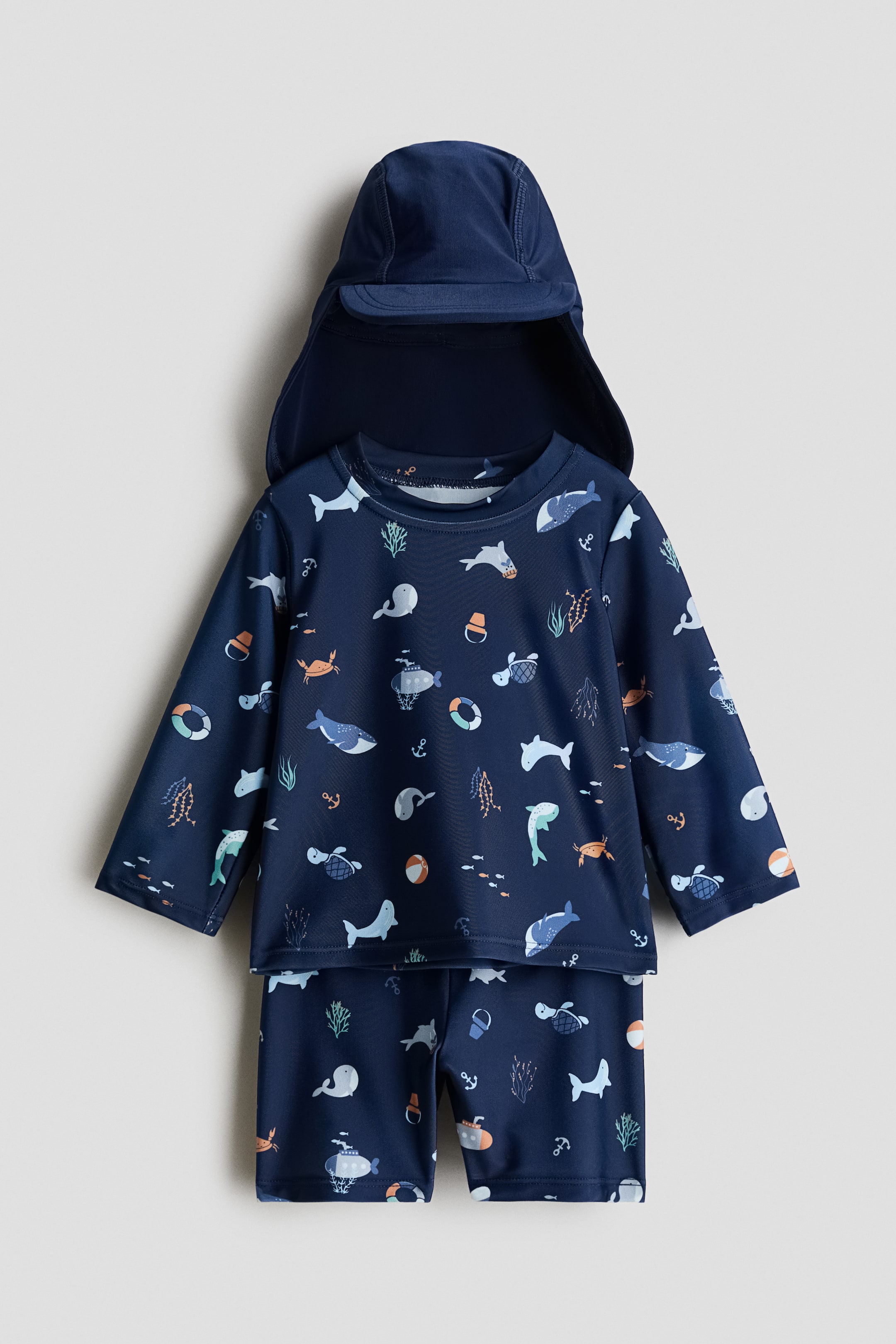 View larger image: A three-piece swim set is shown from the FRONT, featuring a long-sleeved top, shorts, and a cap, all in a dark blue fabric with a colorful sea creature pattern. The top showcases a round neckline and raglan sleeves, the shorts feature an elastic waistband, and the cap includes a front peak with a protective back flap.