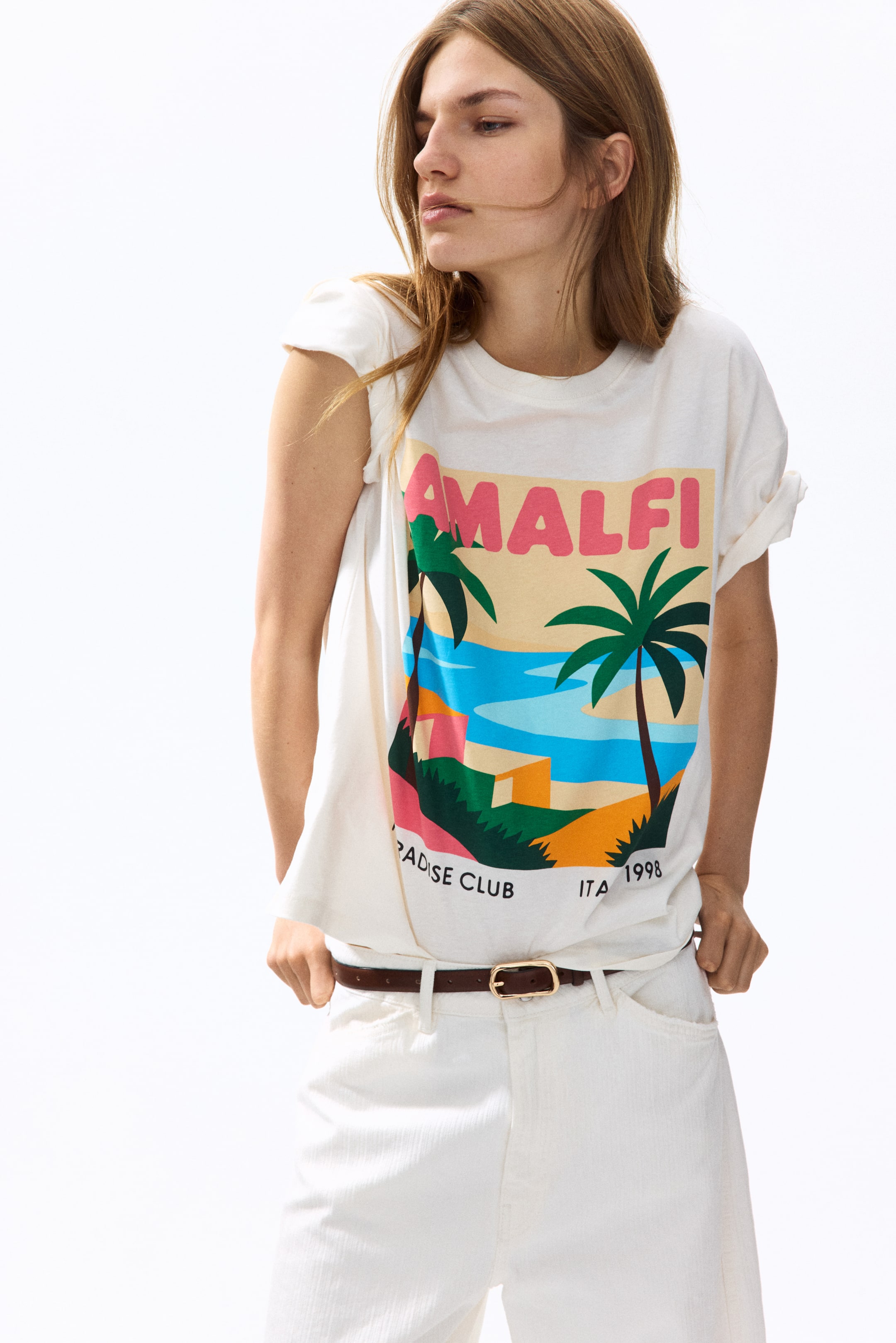 View larger image: A cream t-shirt featuring a colourful graphic print on the front, depicting a coastal scene with palm trees, blue water, and buildings. The graphic includes "AMALFI" in pink text and "PARADISE CLUB ITA 1998" in white text. The short sleeves are rolled up, and the t-shirt appears in a relaxed fit.